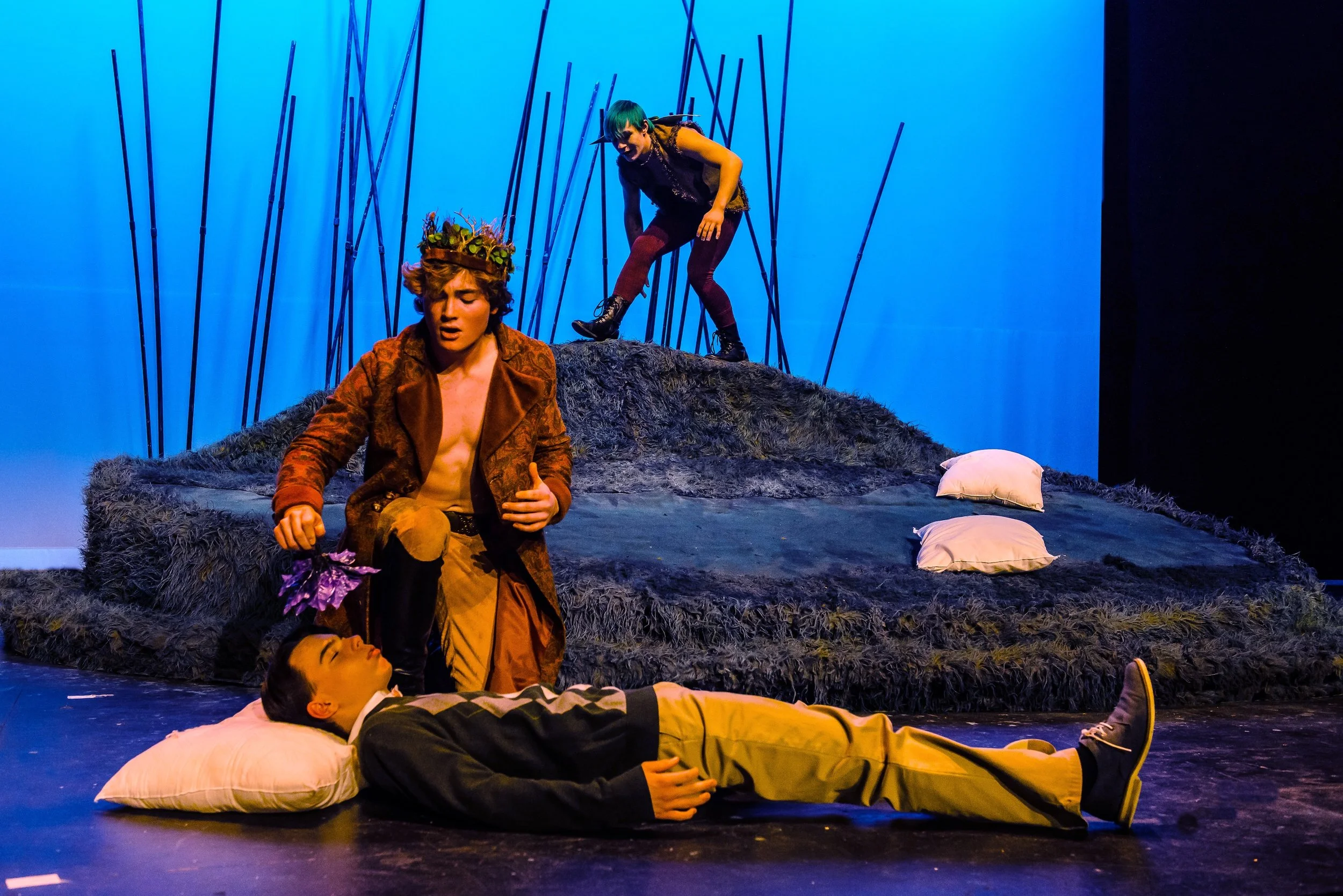 Actors performing on stage in a theatrical play with a blue background, one actor lying down on a pillow and others standing or kneeling nearby.