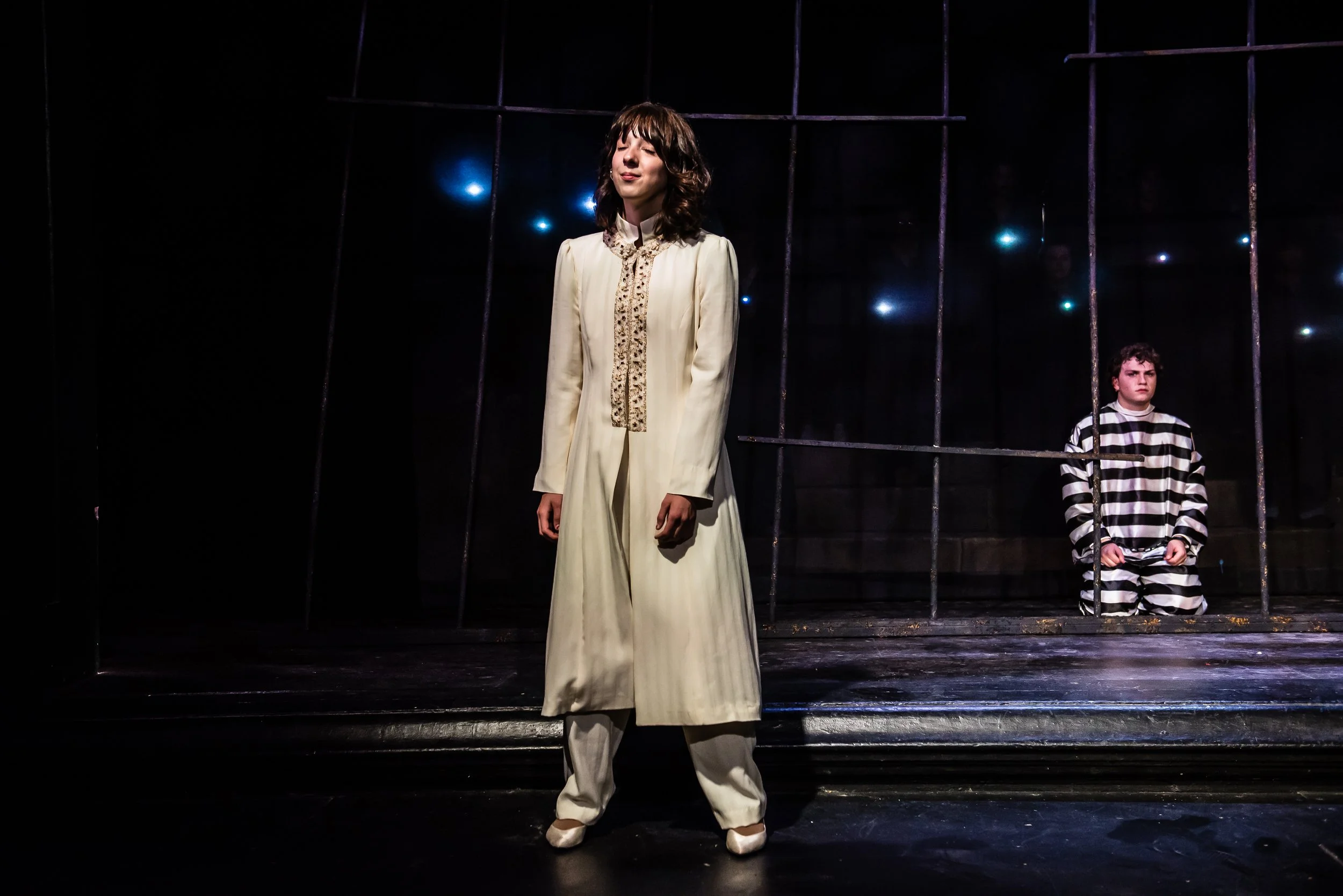 Two actors on stage, one woman in cream-colored vintage dress and shoes, standing in front of a metal cage, and one young man in black-and-white striped pajamas sitting behind bars, with a dark background and faint stage lights.