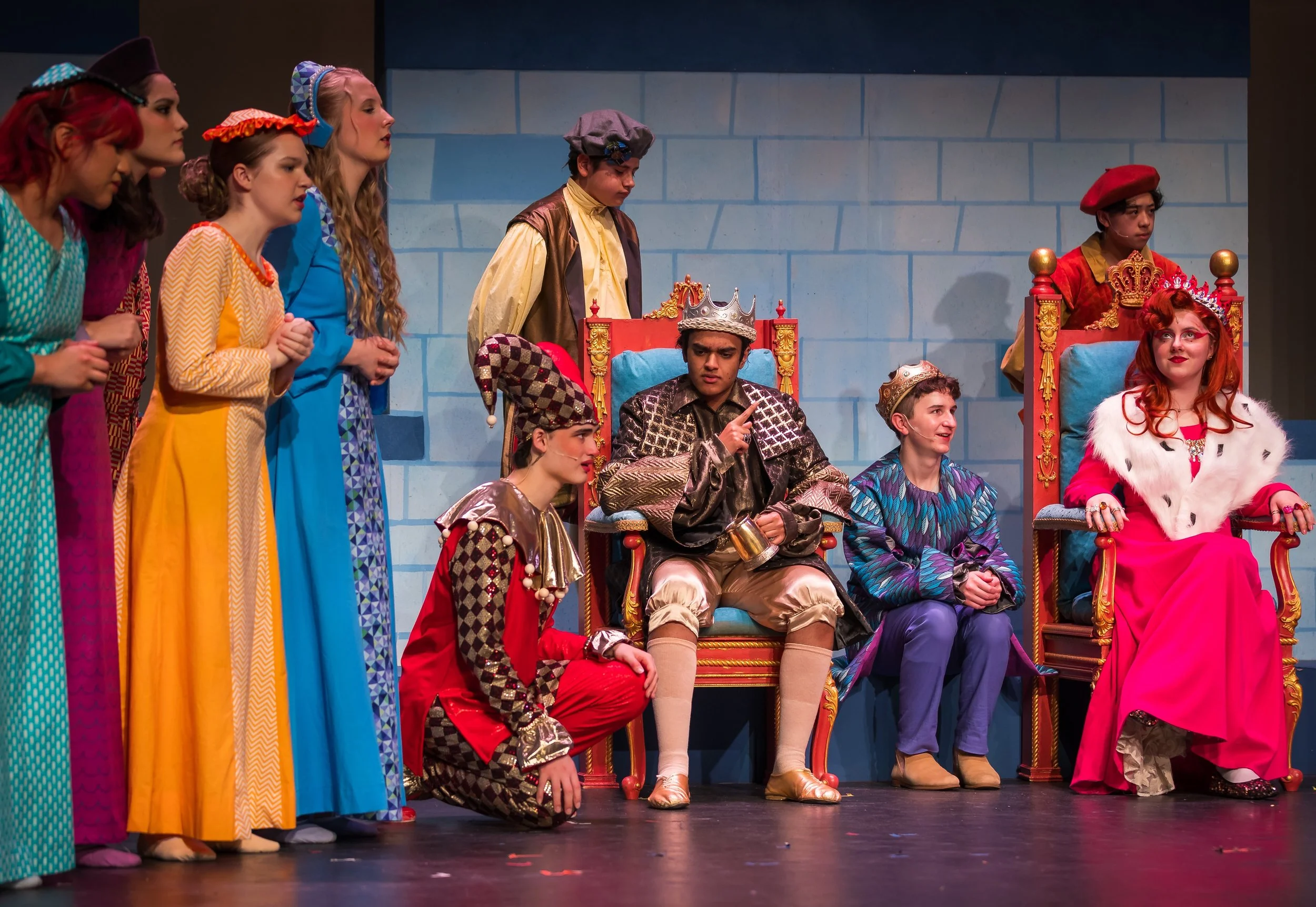 The image depicts a theatrical scene with a group of children dressed in colorful costumes, representing a royal court or castle setting. Some children are sitting on ornate thrones, while others stand beside or behind them engaged in a play, with a 