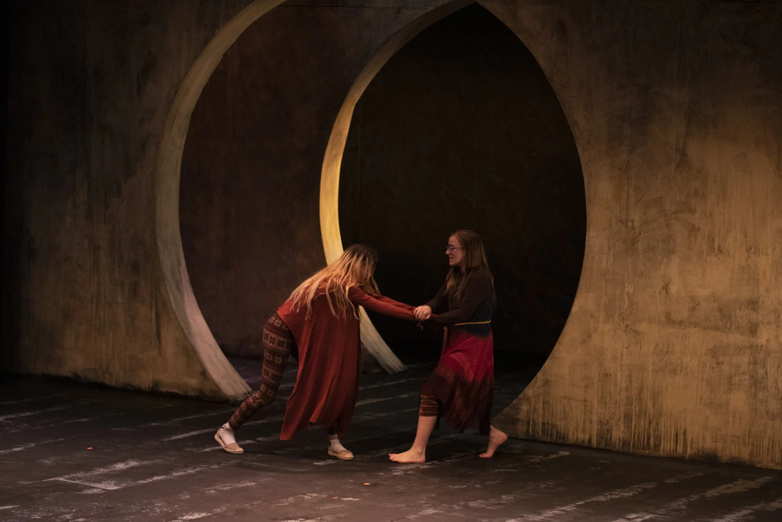 Two women dressed in traditional costumes fight inside a circular opening in a large, textured wall with an arched opening in the background.