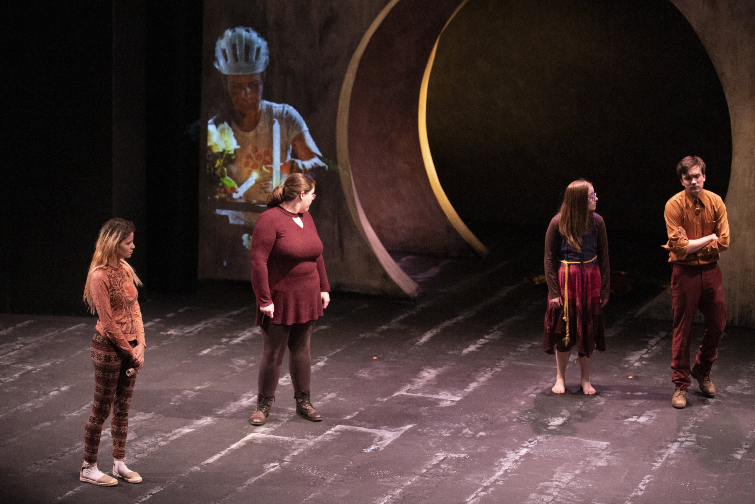 Four performers stand on stage in a theater, with one large screen above showing a close-up of a woman in a helmet and casual clothing.