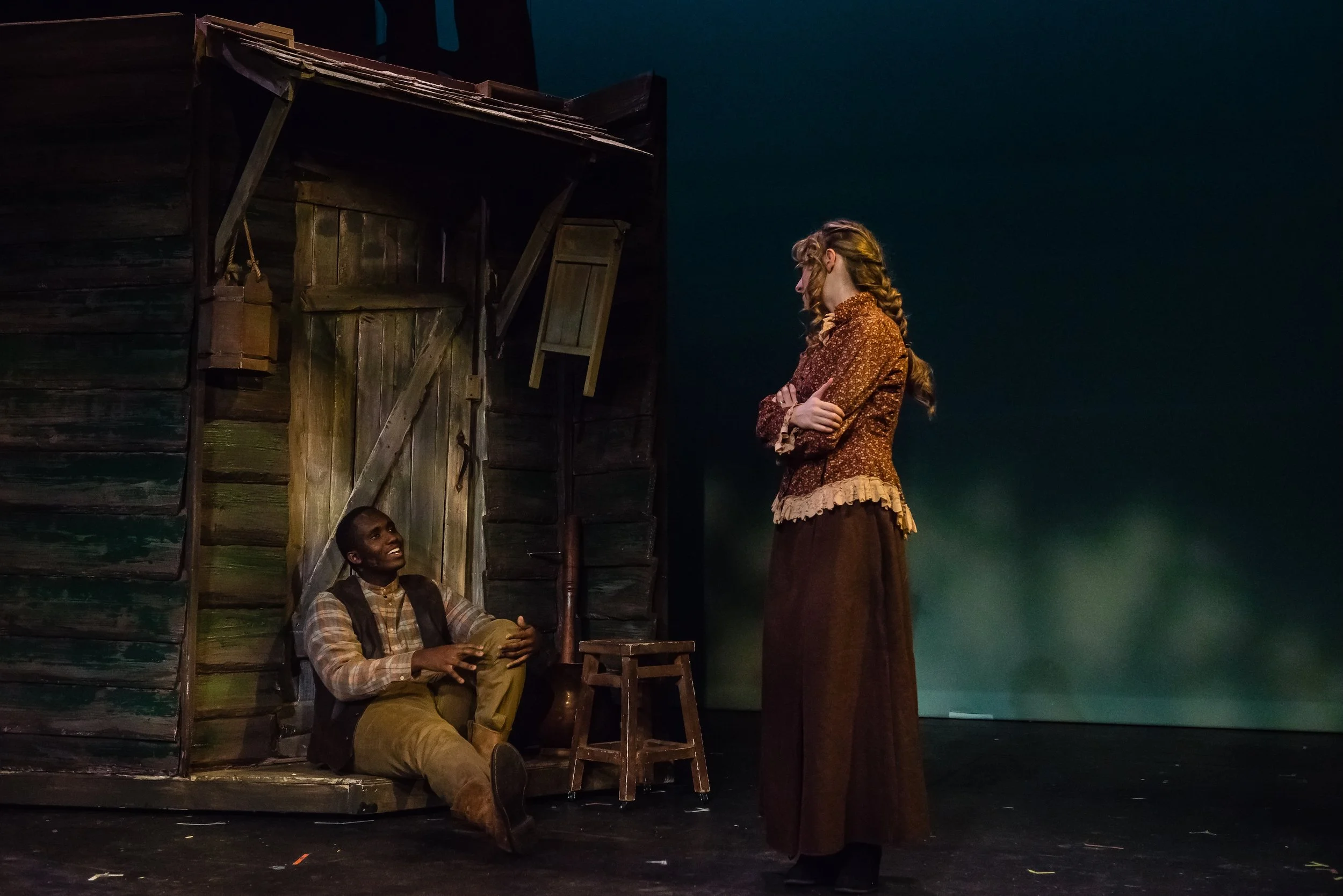 Two actors on stage during a theatrical performance. One with braided hair and a floral dress stands with crossed arms, while the other with short hair and a plaid shirt sits on the floor next to a wooden structure, looking up and smiling.