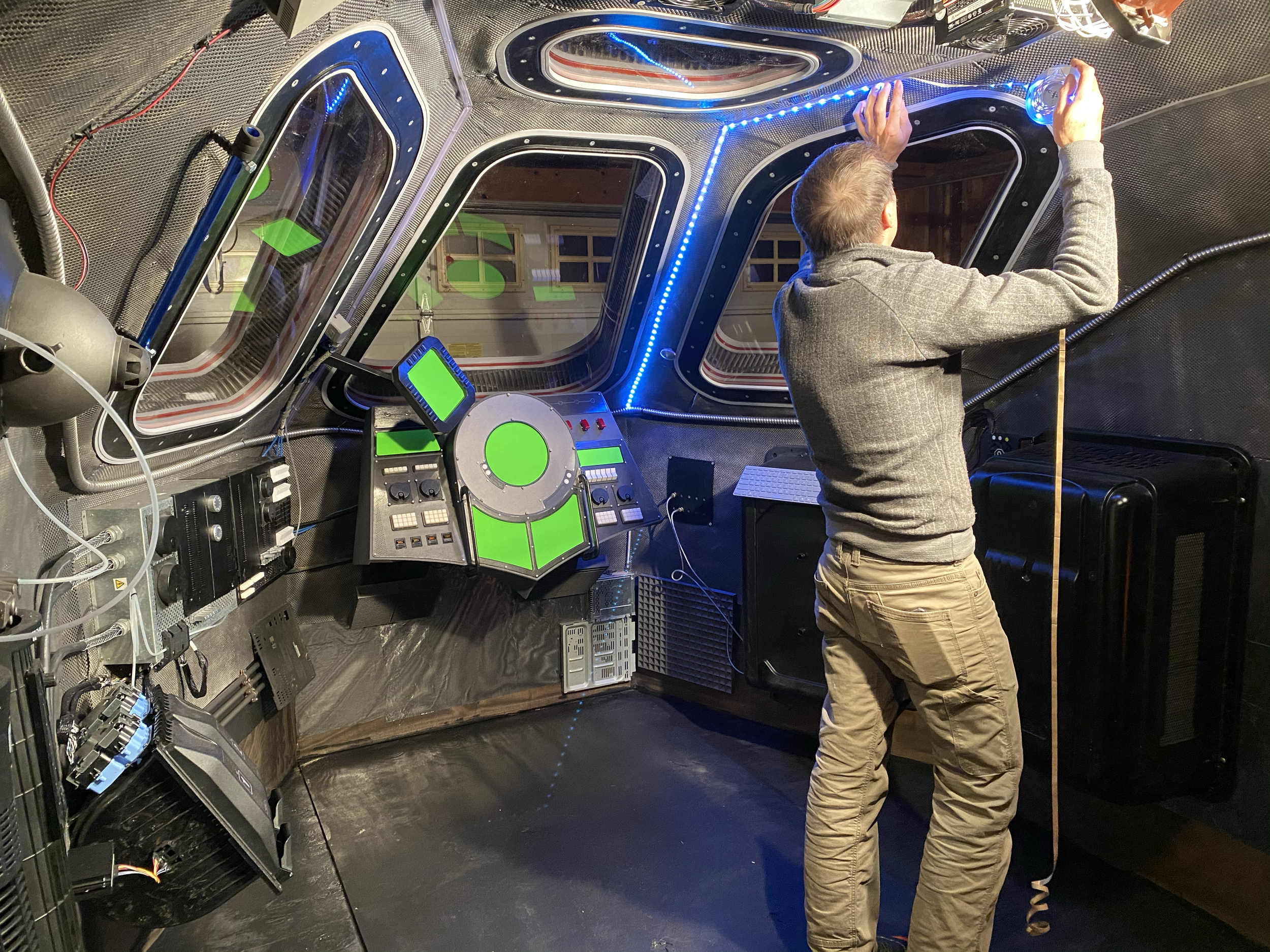 A man working on the interior of a spaceship film set, surrounded by electronic control panels, tubes, wiring, and windows with a dark background.