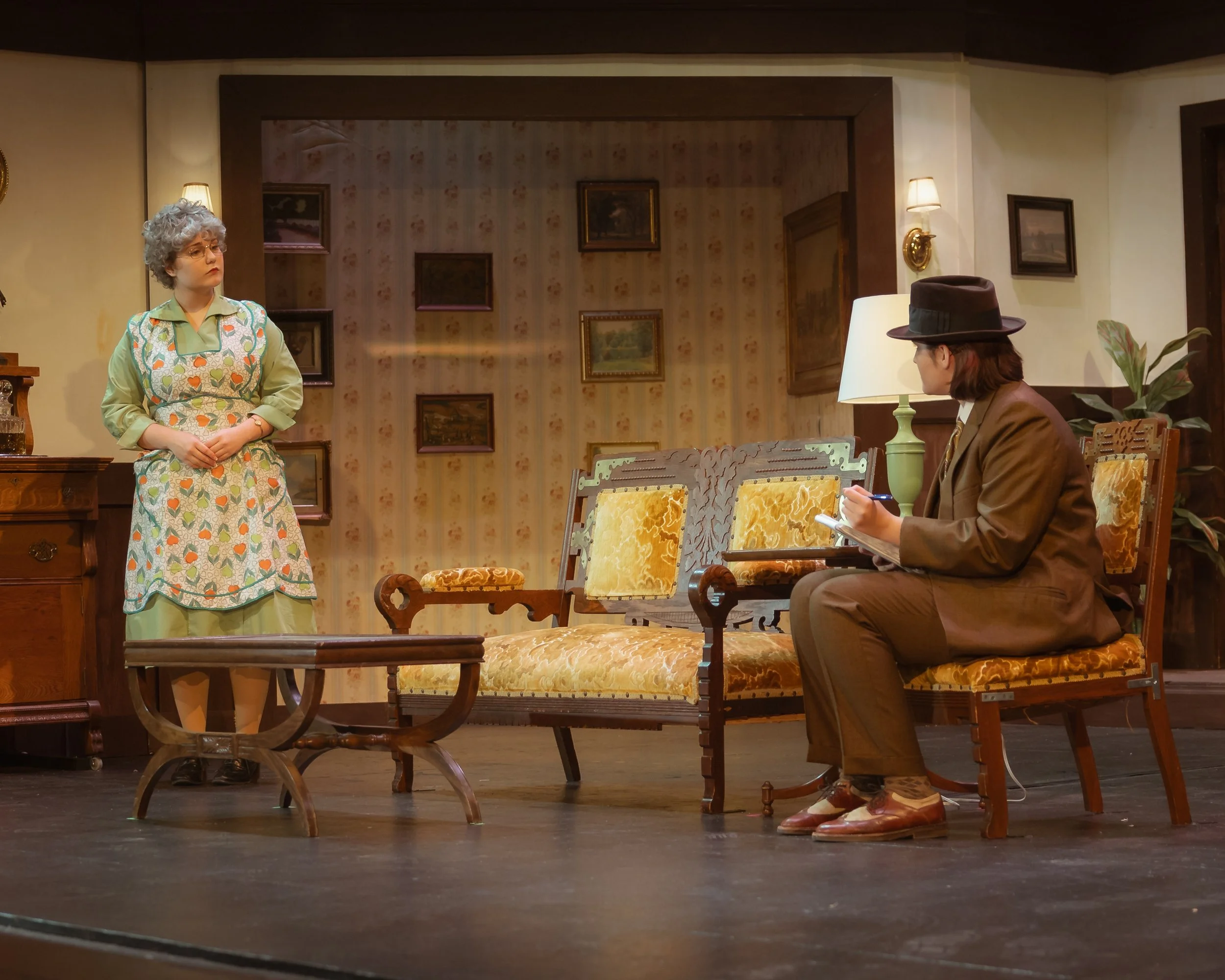 Two people on stage in a living room setting, one woman standing and one man sitting on a bench. The woman has curly gray hair, glasses, and a colorful dress with a heart and pear pattern. The man wears glasses, a brown jacket, brown hat, and tan sho