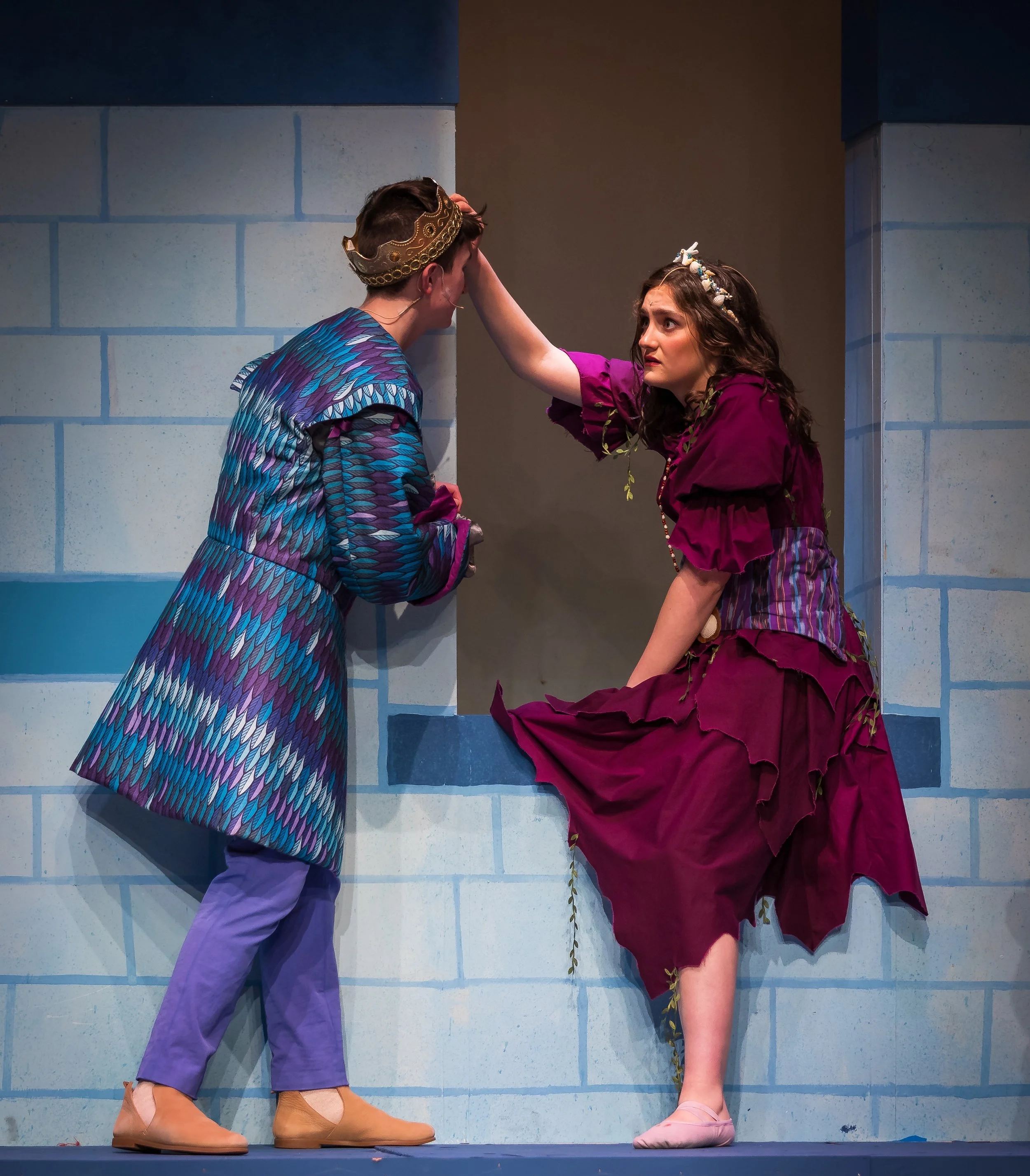 Two performers on stage in a theatrical production, one standing and one balancing on one foot with her leg against a wall, both wearing colorful costumes and crowns, set against a blue tiled wall background.