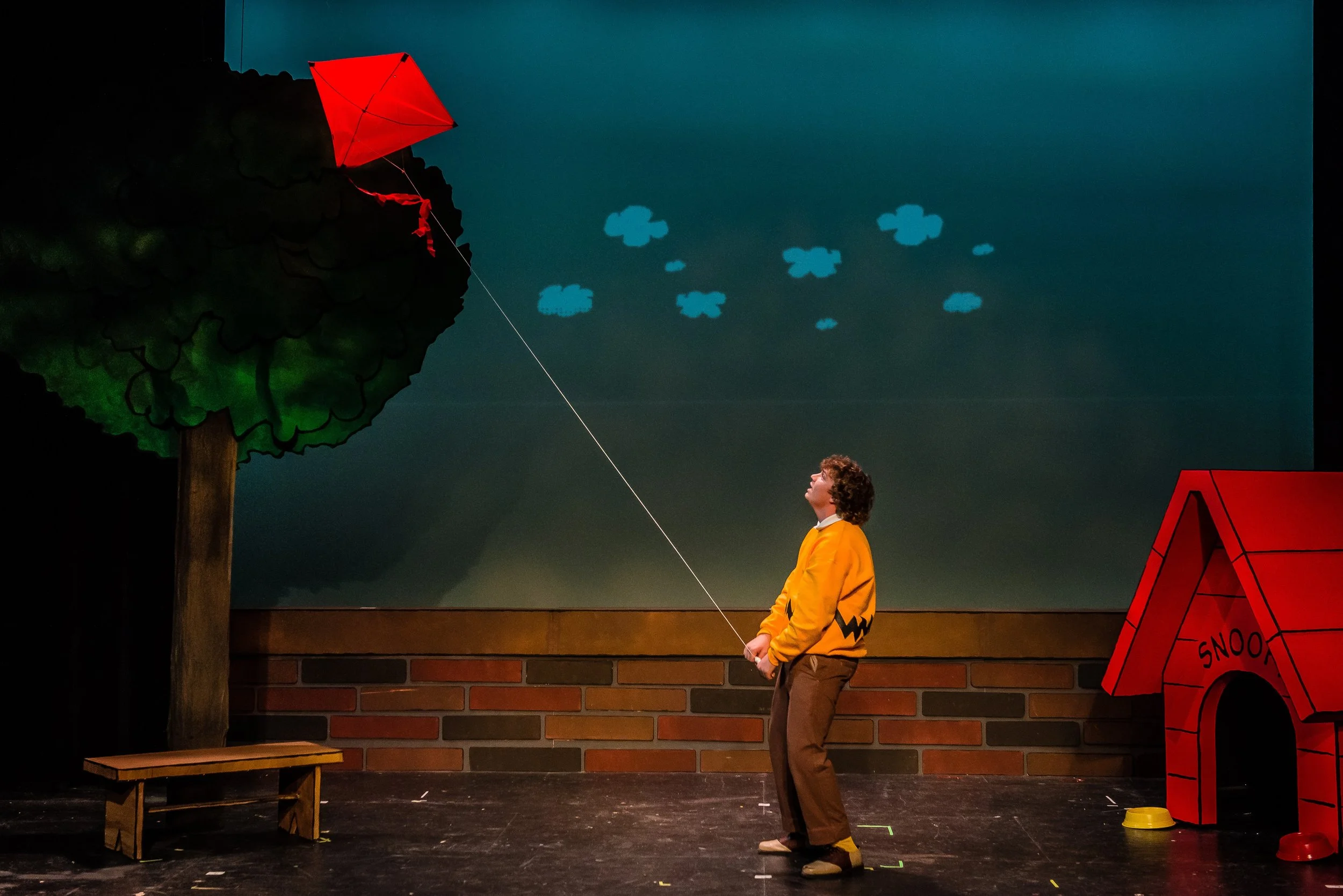 An actor playing Charlie Brown is on stage flying a red kite. The stage background is blue with small clouds, and there is a cartoon-style doghouse labeled 'Snoopy' on the right and a cartoon tree on the left.