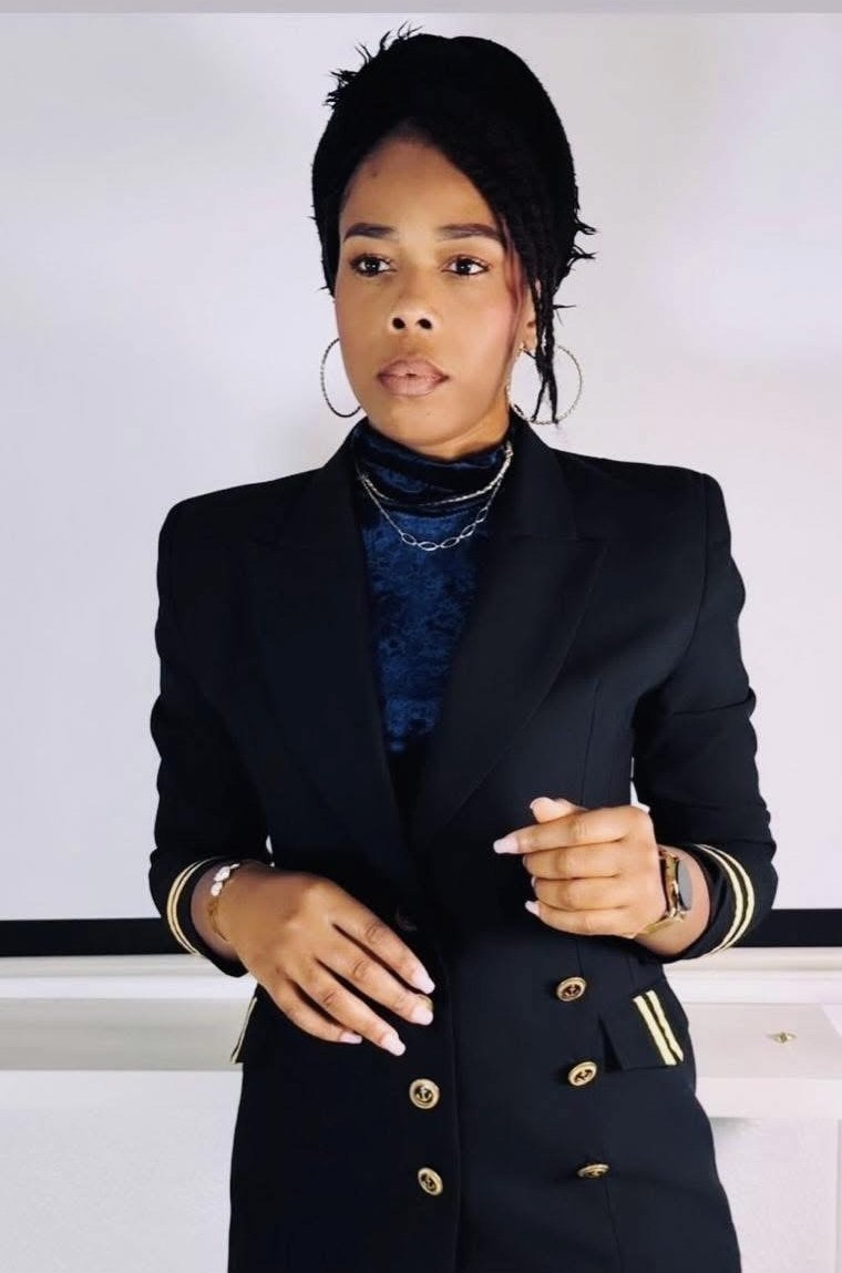 Une femme noire en costume noir avec des boutons dorés, portant des boucles d'oreilles en anneau, des colliers, une montre et un bracelet. Elle a des cheveux courts et noirs, et elle pose devant un mur blanc.