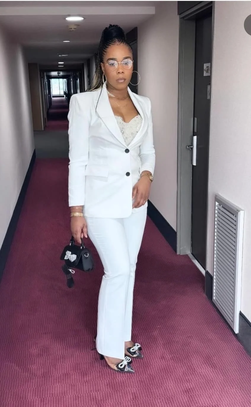 Femme élégante en costume blanc, portant des lunettes et des bijoux, dans un couloir d'hôtel.