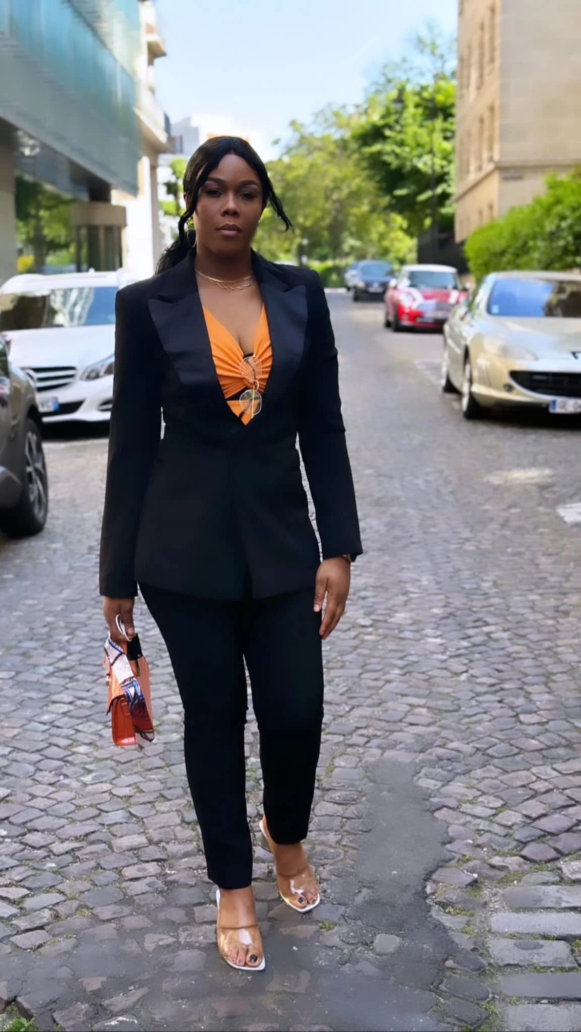 Femme marchant sur une rue pavée, portant un costume noir avec un haut orange, des talons transparents et tenant un portefeuille assorti.