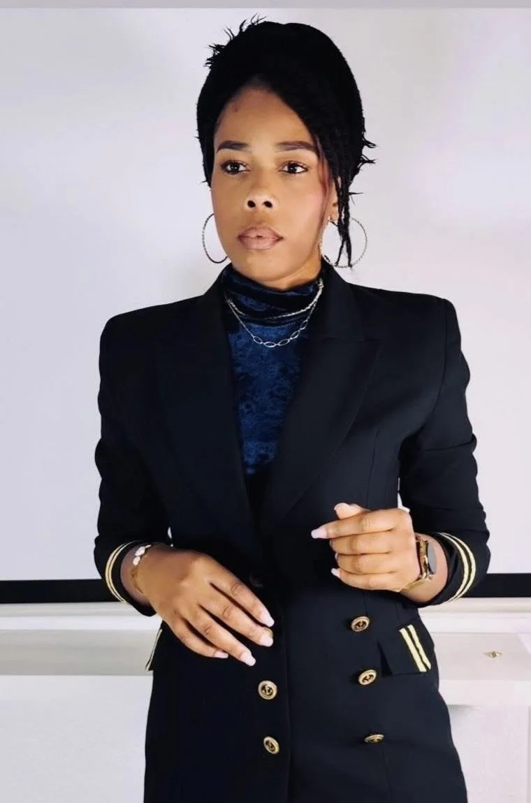 Une femme noire habillée d'un costume noir avec des détails dorés, portant des boucles d'oreilles à cercle, un col roulé en velours bleu, des colliers et une montre, debout devant un fond blanc.