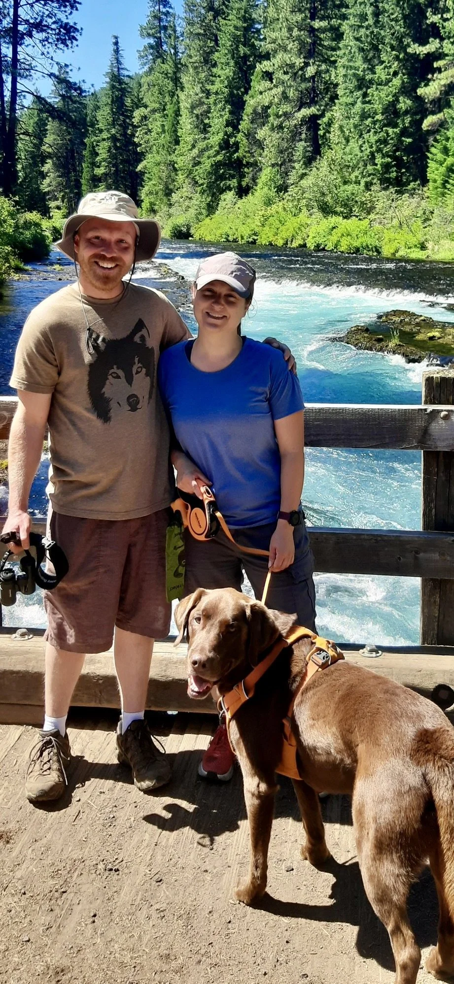 Two people standing on a wooden bridge with a river and lush green forest in the background. One person is holding a brown dog on a leash.