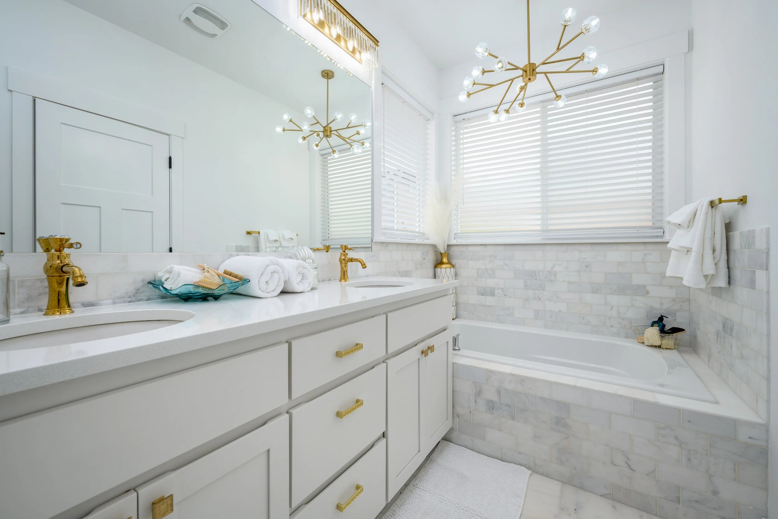 A bright bathroom with white walls, a marble countertop, and gold fixtures. There are white towels, a blue soap dish, and decorative items on the counter. A large mirror reflects two modern chandeliers with multiple bulbs, and there is a window with 