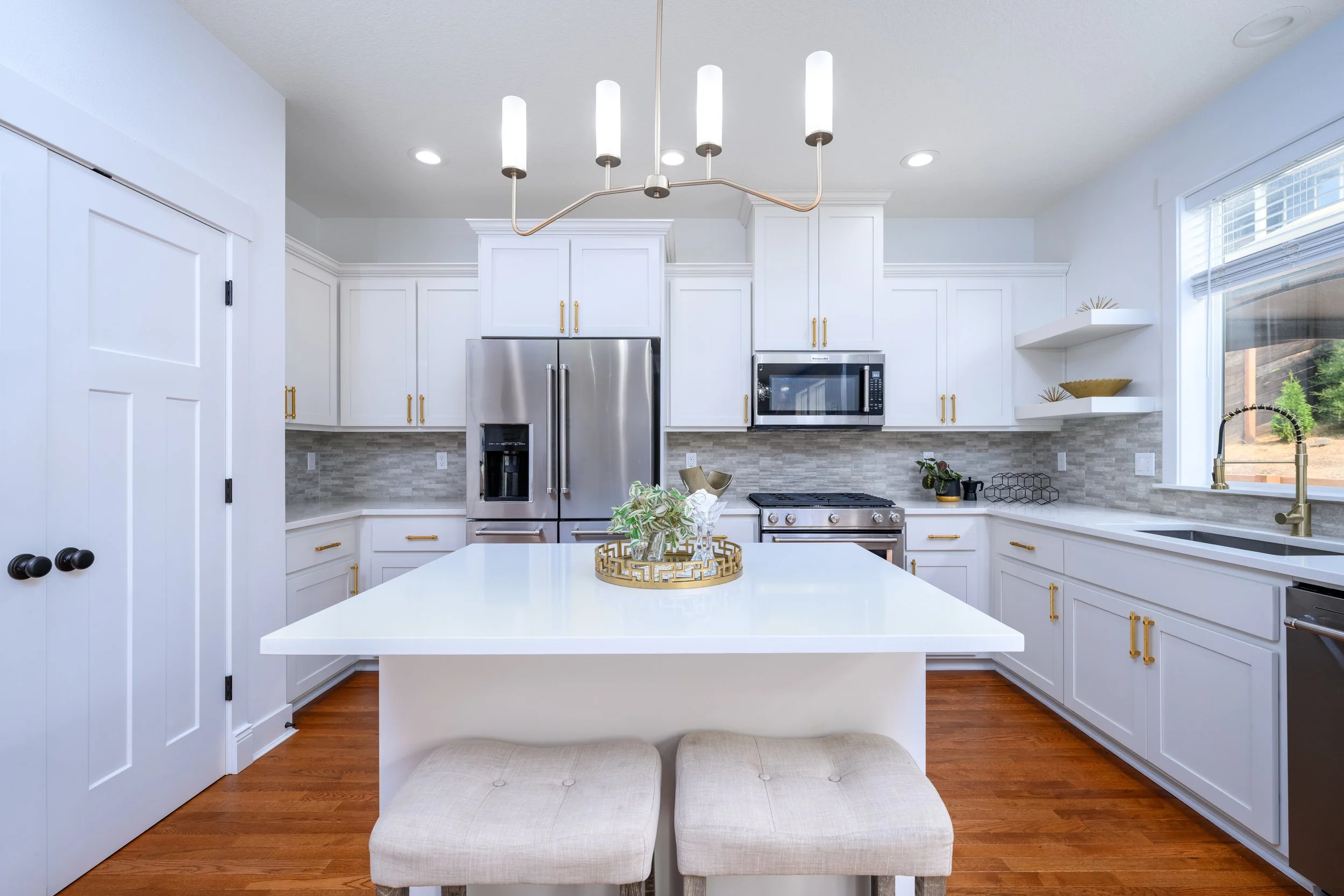 Modern kitchen with white cabinets, stainless steel refrigerator, microwave, and oven, wooden floor, and a white island counter with a decorative tray and plant, sunlit from large window.