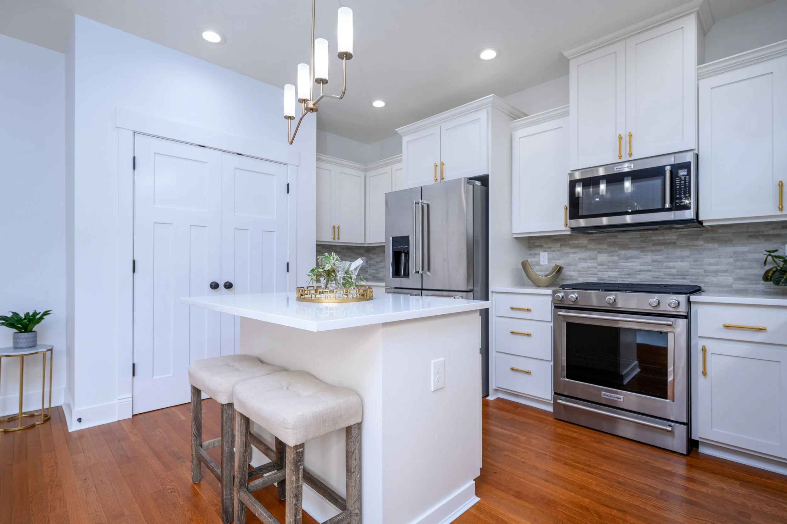 Modern kitchen with white cabinetry, stainless steel appliances, pendant light fixture, small beige upholstered stools, decorative items on counter, hardwood floors, and a plant on a side table.