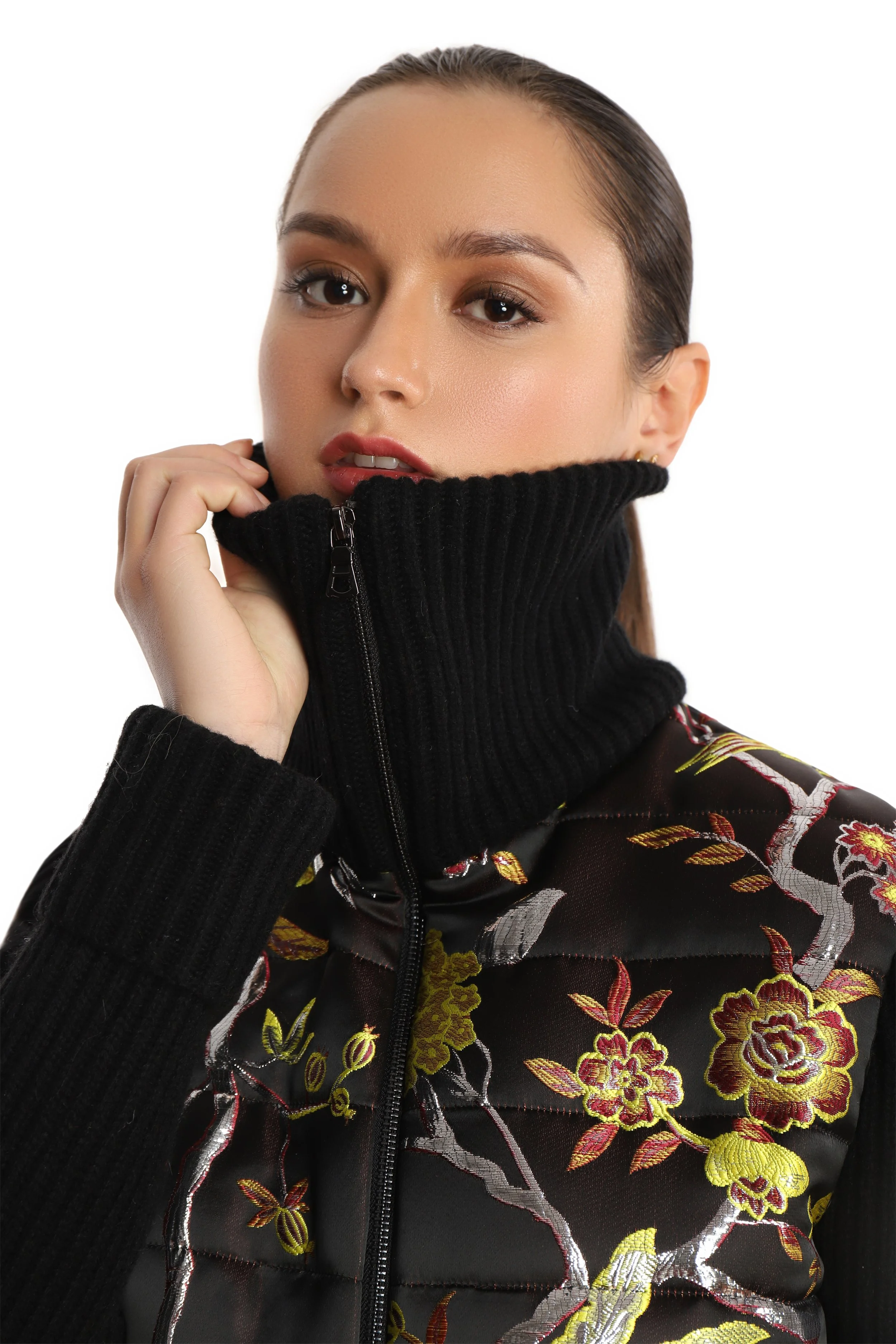 Close up of model wearing a multi colored embroidered floral baseball jacket with black rib knit sleeves and high neck rib knit collar. 