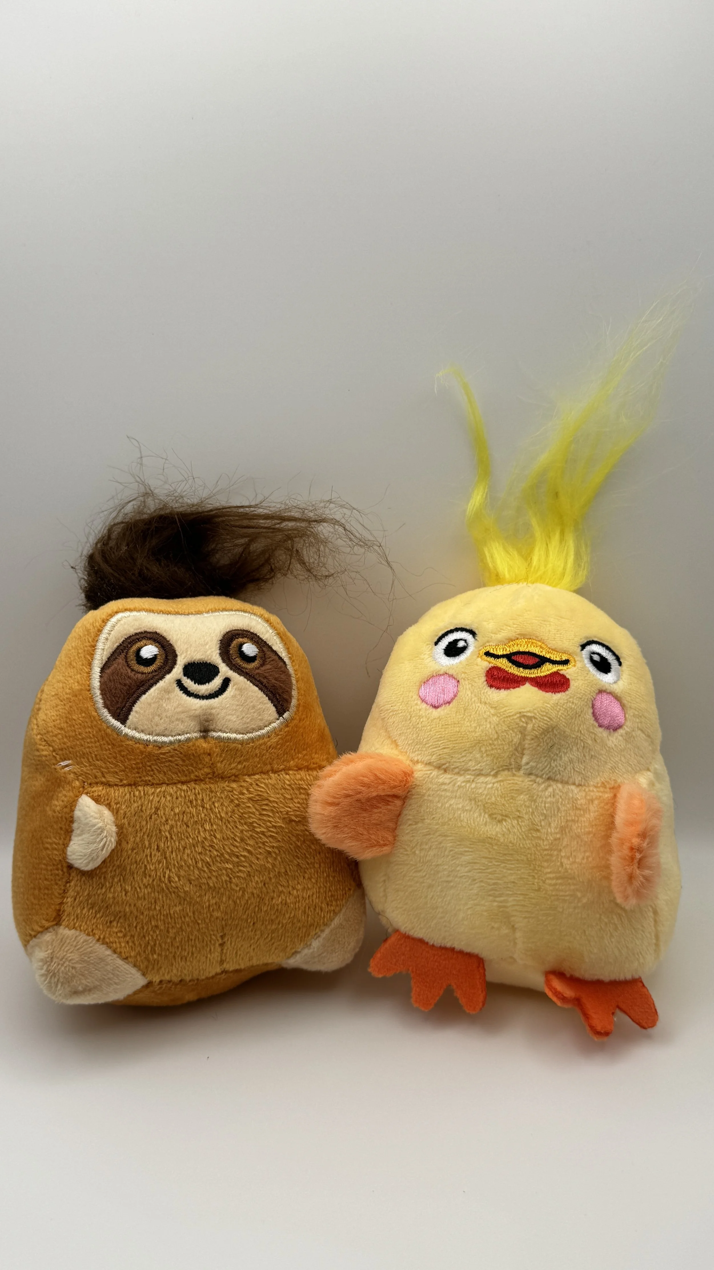 Two plush toys resembling a sloth and a chick with a chick's face, yellow feathers, pink cheeks, orange feet, and a tuft of yellow hair, placed on a white surface against a plain background.
