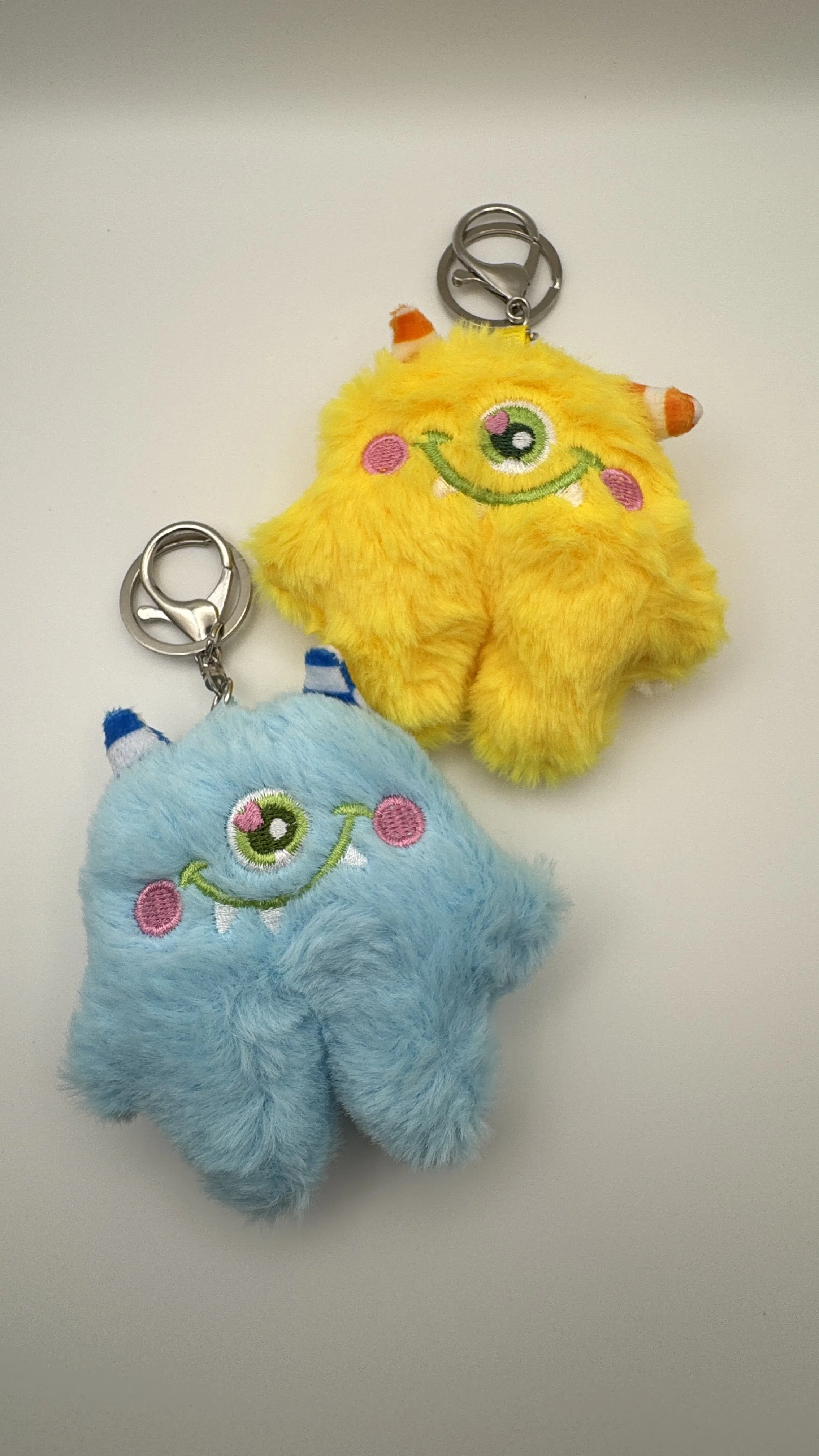 Two plush monster-shaped keychains, one yellow and one blue, with embroidered smiling faces and single large eyes, each with small horns and a metal keyring attached.