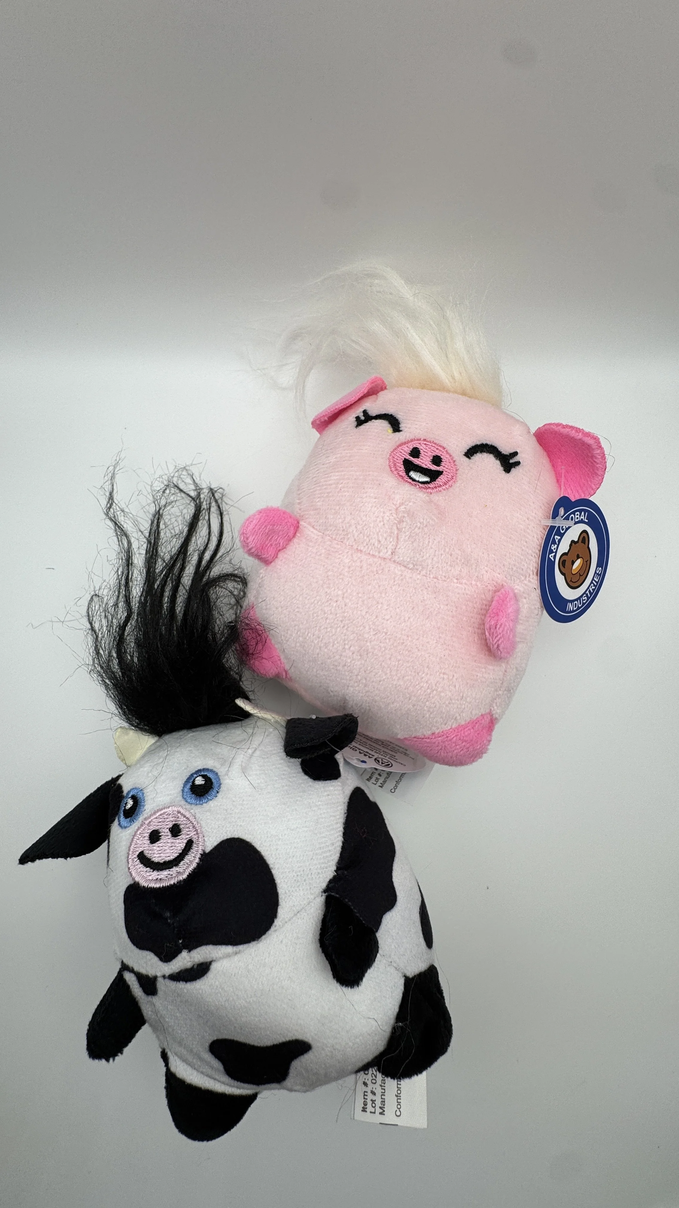 Two plush pig toys, one pink with a smiling face and blonde hair, the other black and white cow with a smiling face and blue eyes.