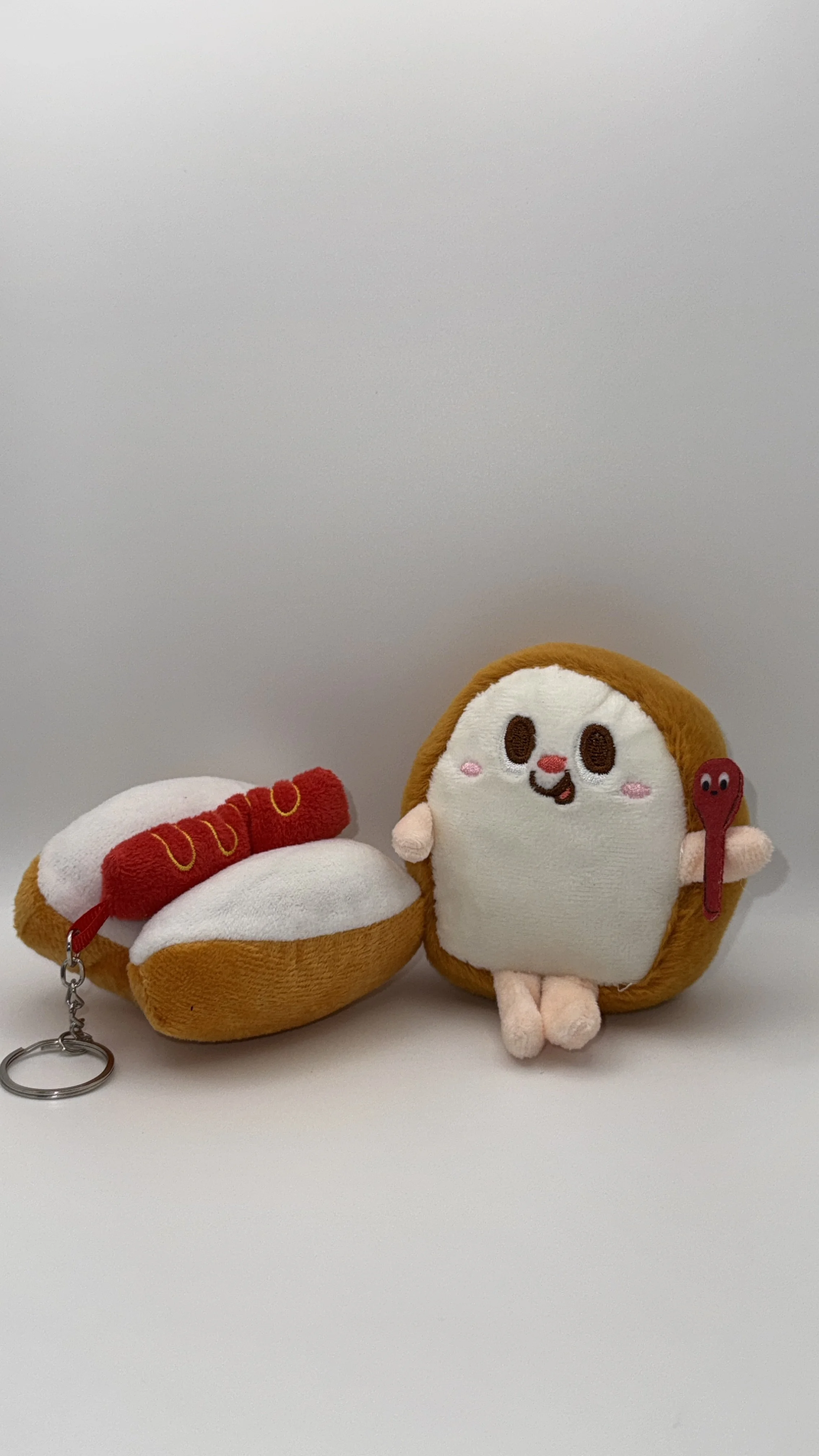 A plush toy of a slice of bread with a happy face, pink cheeks, and little arms and legs, holding a wooden spoon, next to a plush toy of a hot dog with a smiling face and yellow loops, on a white background.