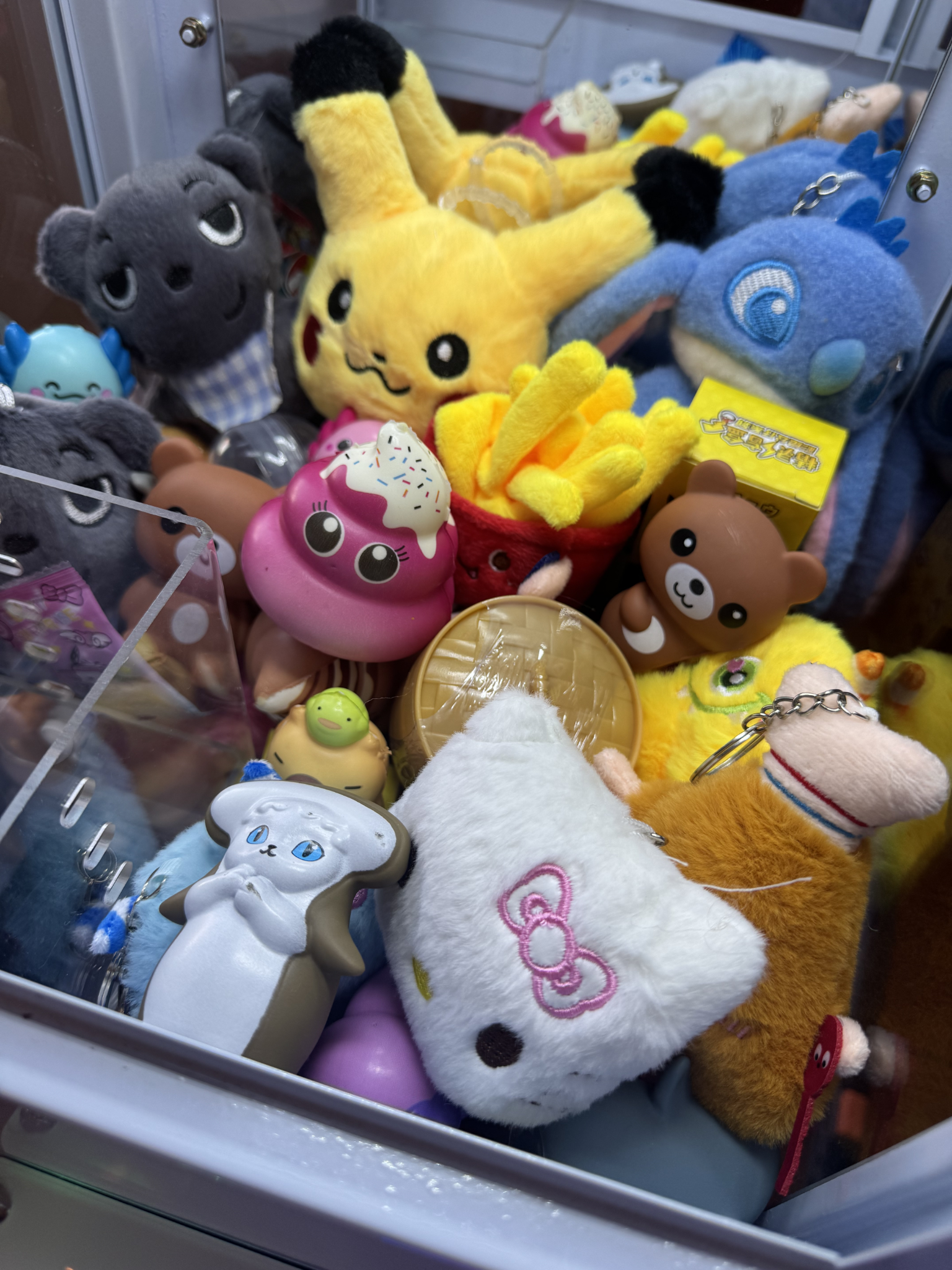 A claw machine filled with various plush toys including a Pikachu plush, squishies, a Hello Kitty prizes, blind boxes, and other colorful stuffed animals.