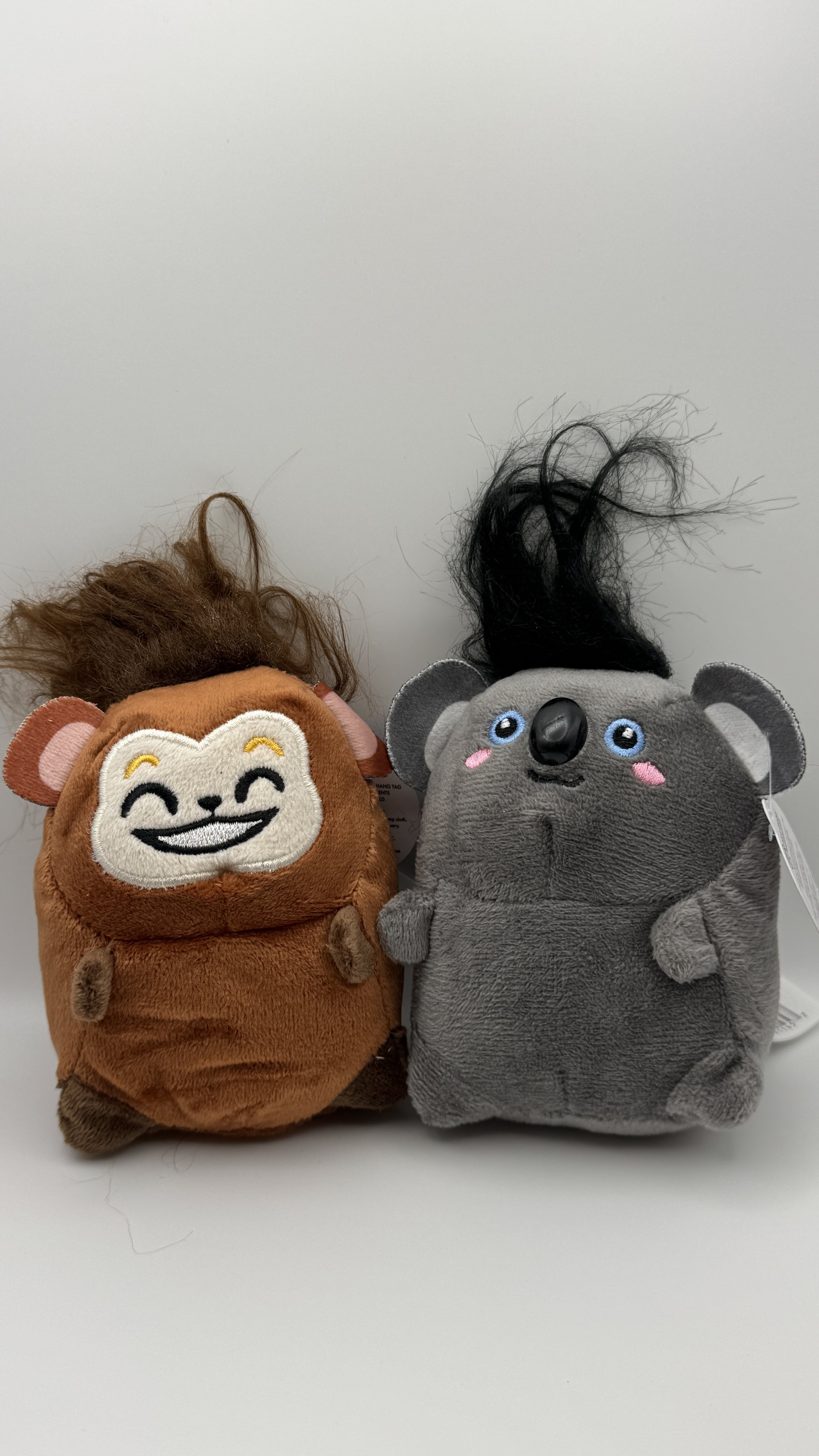 Two plush animal toys, a brown lion with a happy face and a gray koala with pink cheeks, blue eyes, and black hair, against a plain white background.