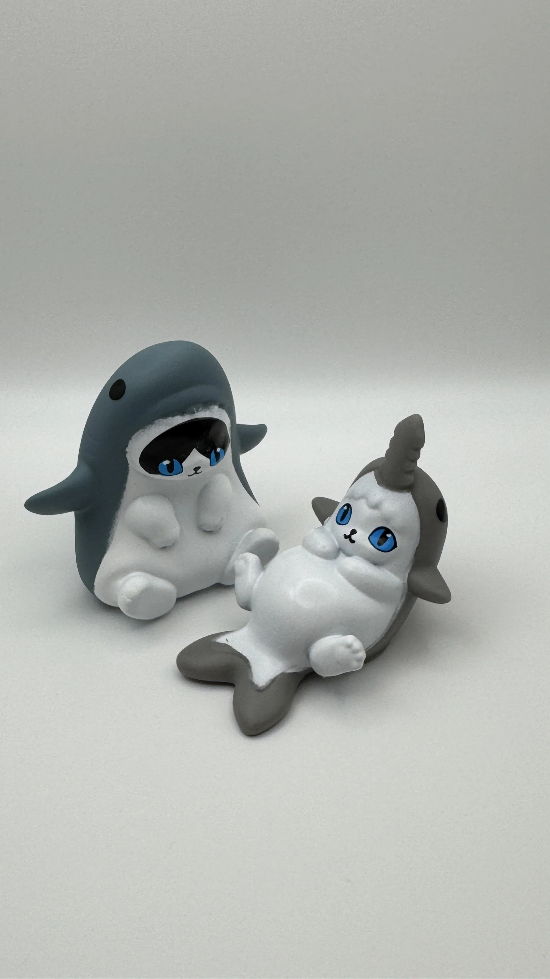 Two plush toys resembling cute animals, one dressed as a shark and the other as a cat, lying on a white surface.