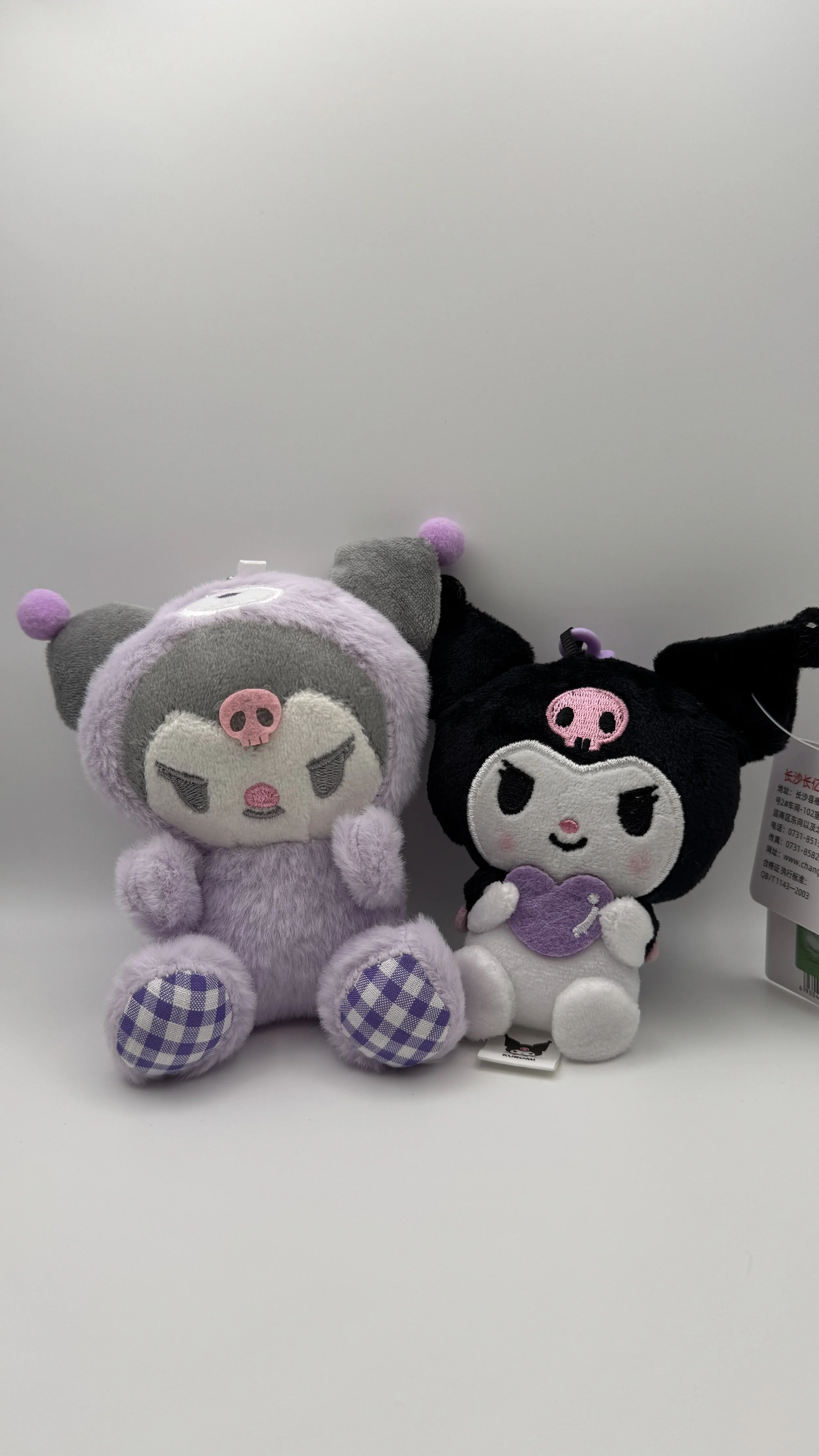 Two plush toys with cartoonish faces and skull motifs on their heads, one purple and one black, sitting on a white surface against a plain white background.