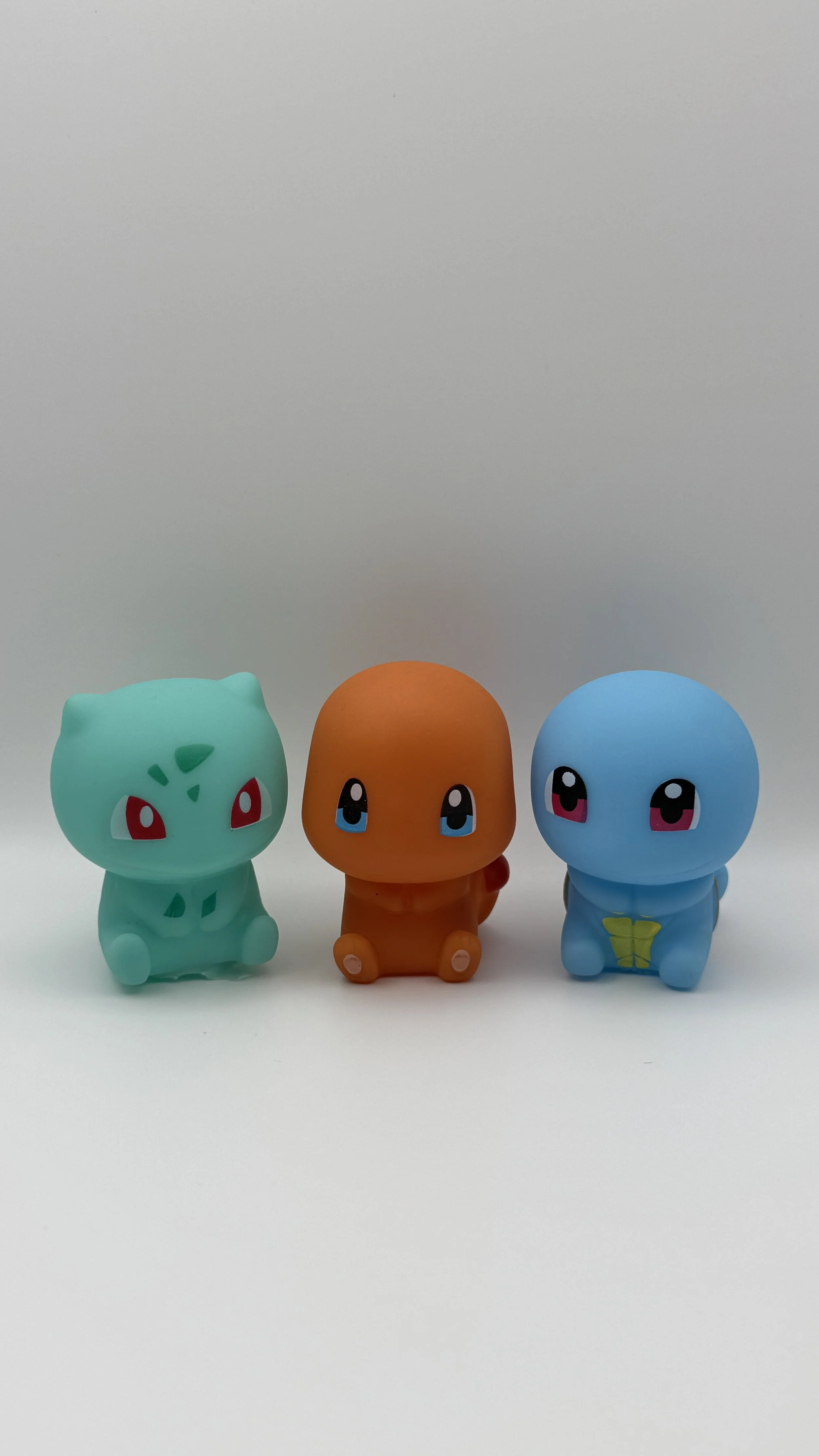 Three Pokémon figures, Bulbasaur with green body and red eyes on the left, Charmander with orange body and blue eyes in the middle, Squirtle with blue body and pink eyes on the right, are displayed on a white surface against a plain background.