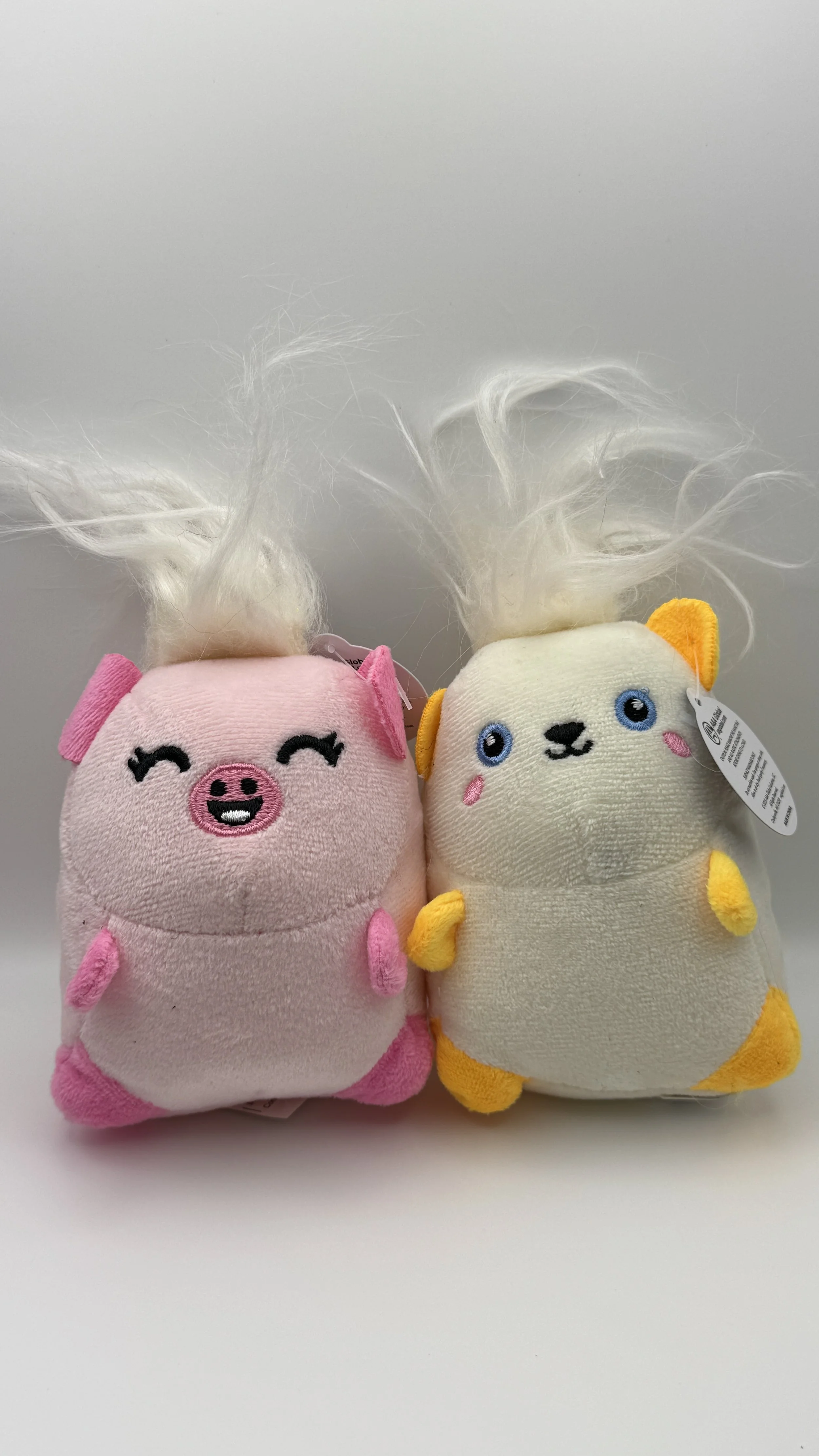 Two plush toys resembling cute animals with fluffy hair, one pink pig and one cream-colored dog with yellow accents, positioned side by side on a white surface.