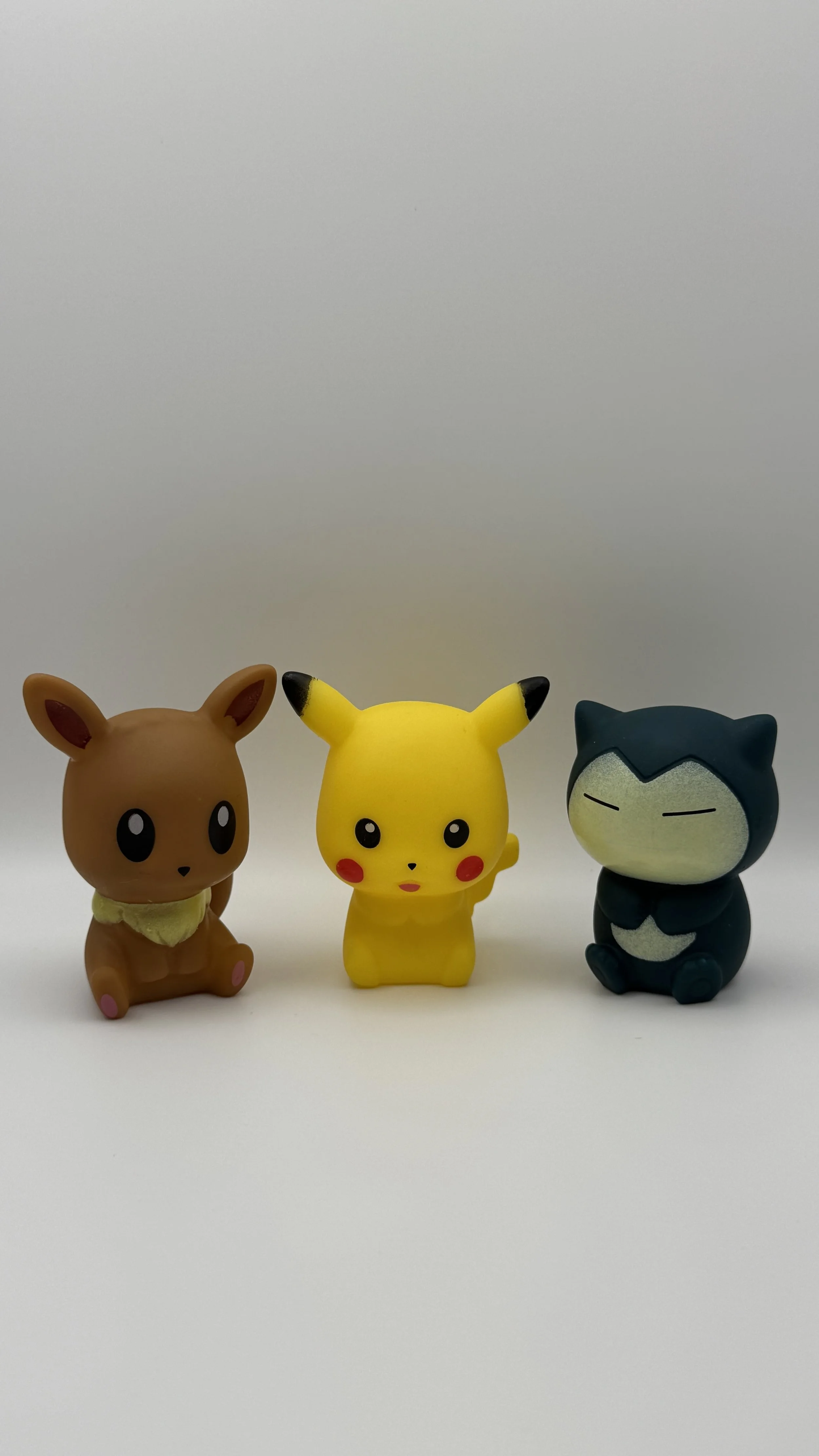 Set of three small Pokémon figurines on a white surface with a plain background. The figurines include Eevee on the left, Pikachu in the middle, and Snorlax on the right.