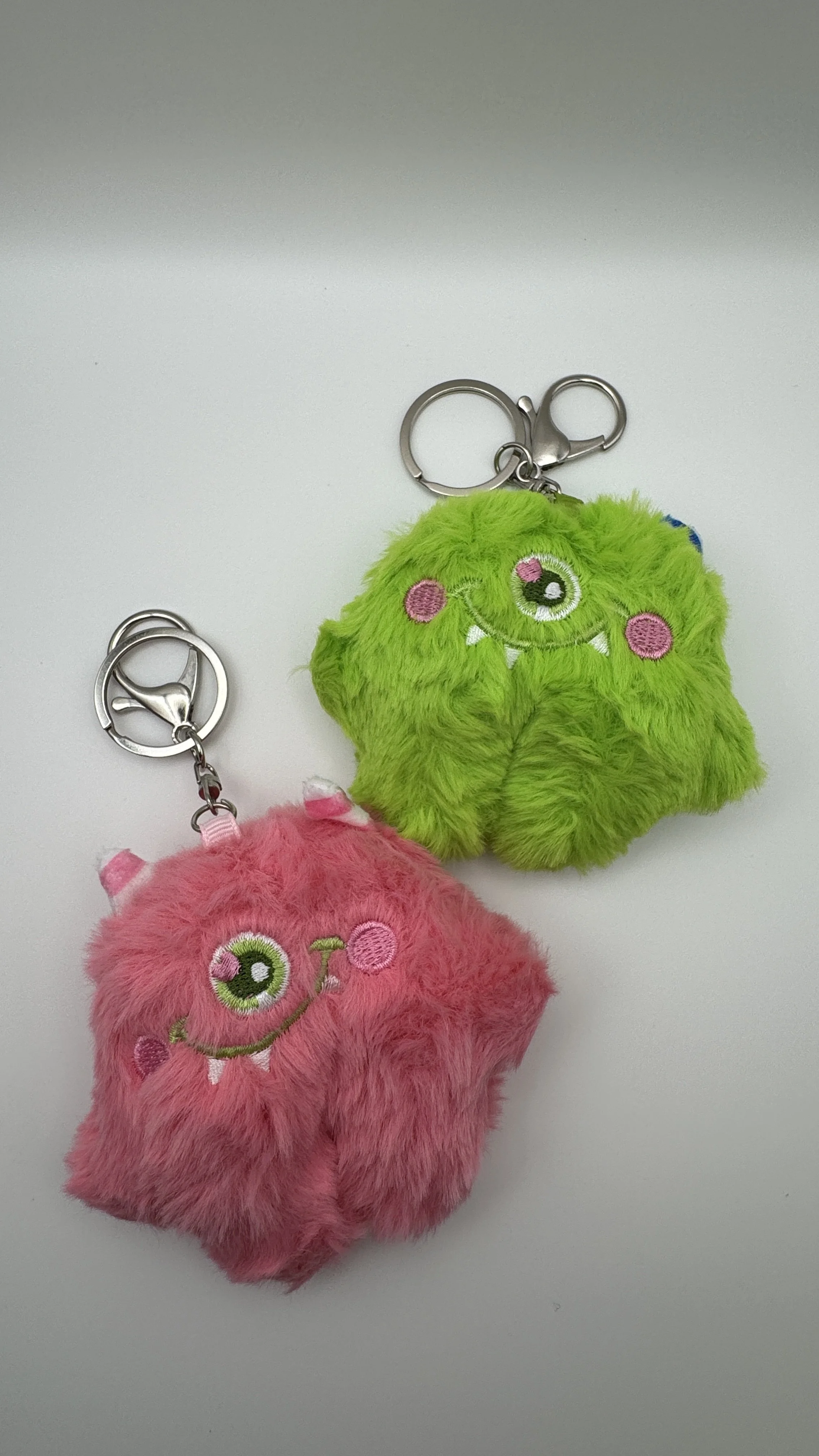 Two fluffy plush keychains, one pink and one green, with embroidered smiling monster faces, on a plain white surface.