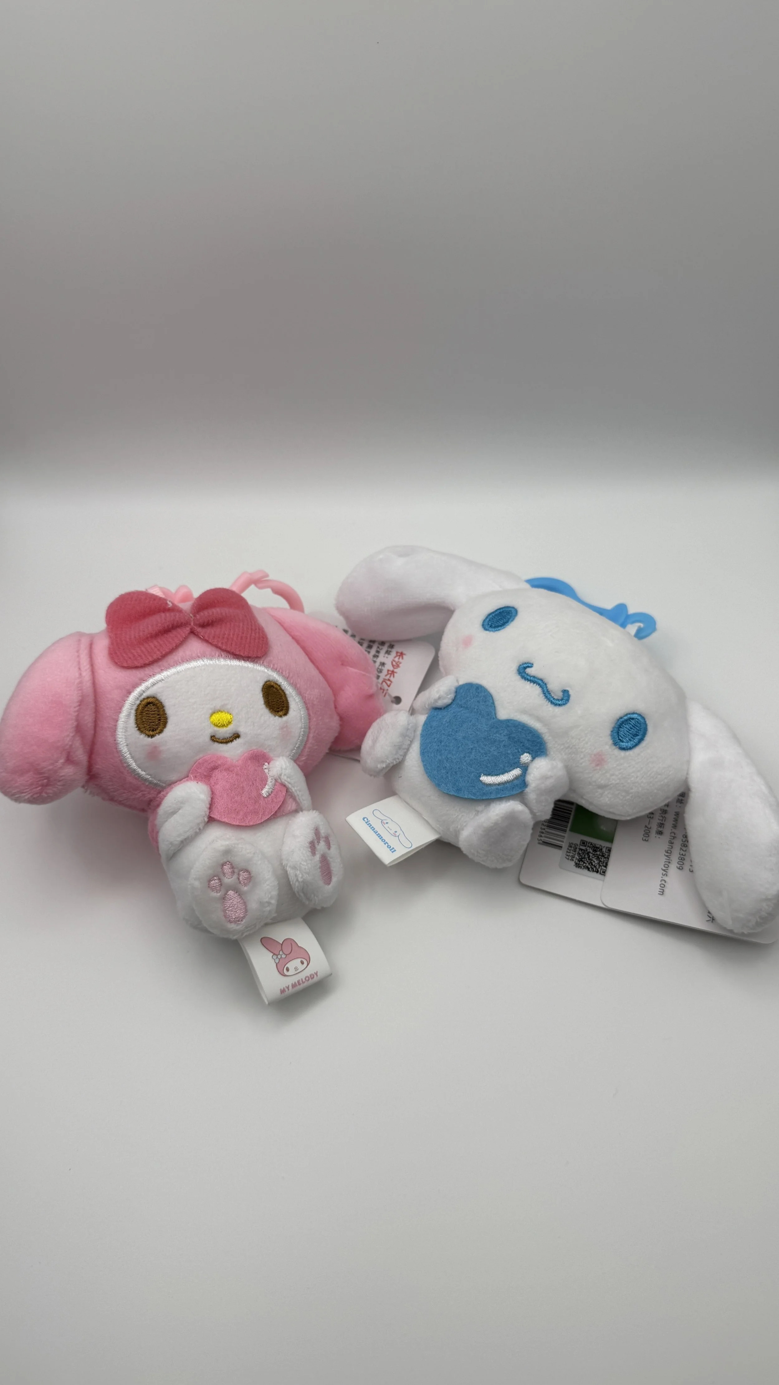 Two small plush toys featuring My Melody characters from Sanrio, one pink and the other white and blue, lying on a white surface against a gray background.