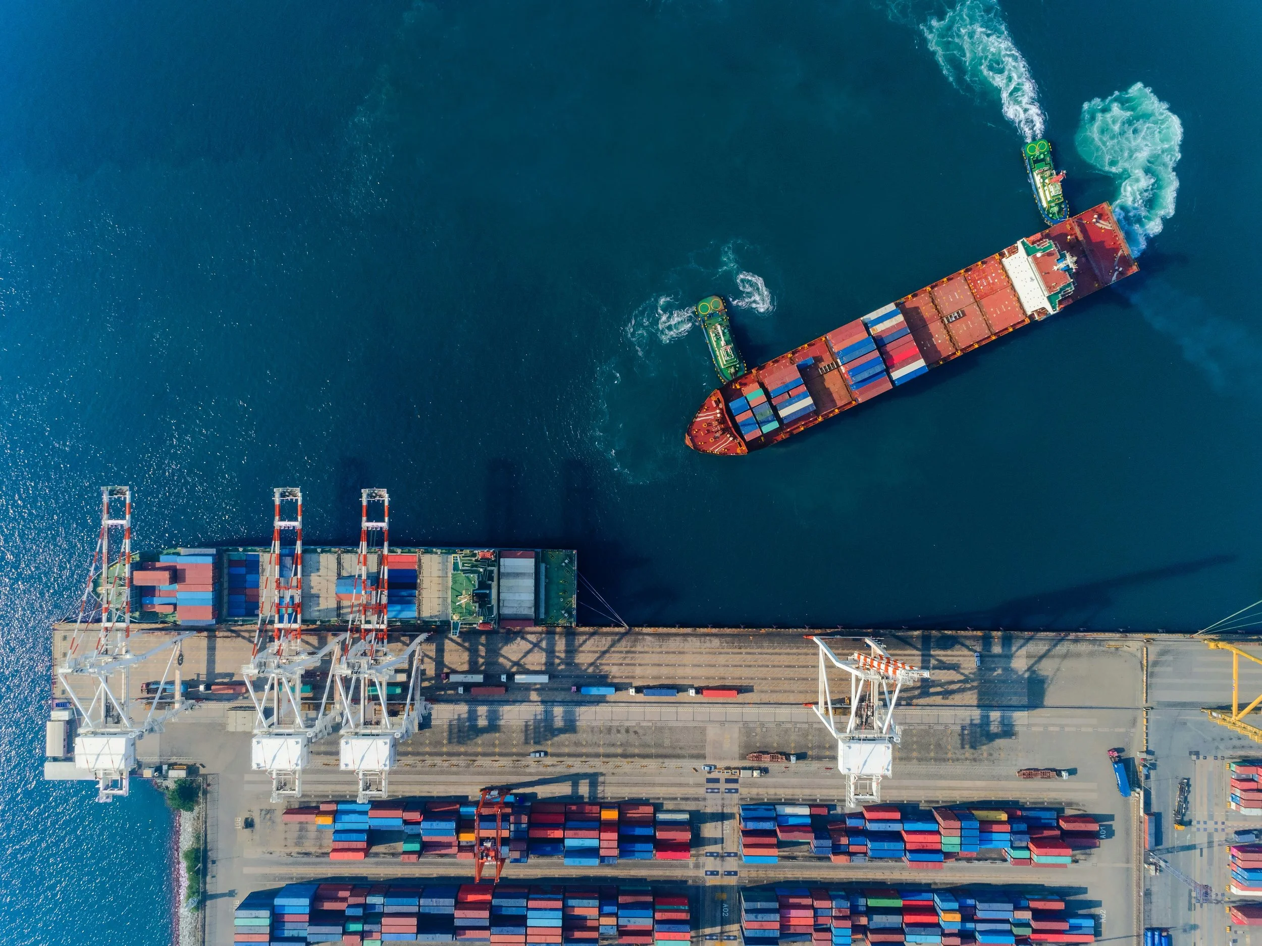 An aerial view of a cargo ship navigating away from a port with multiple cranes and numerous containers on the dock, on a bright day.
