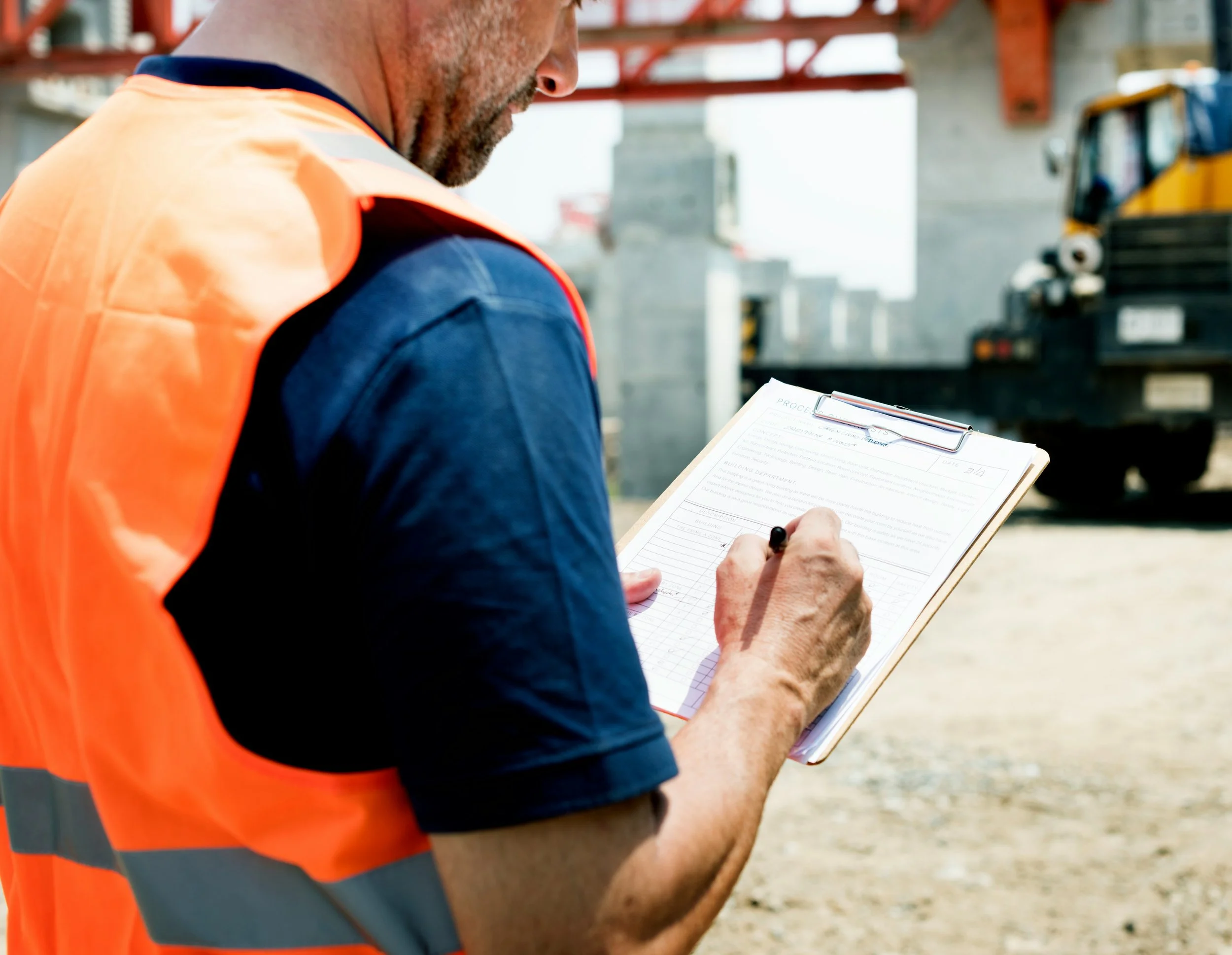 A construction worker in a neon orange safety vest writing on a clipboard at a construction site with steel beams and a truck in the background.
