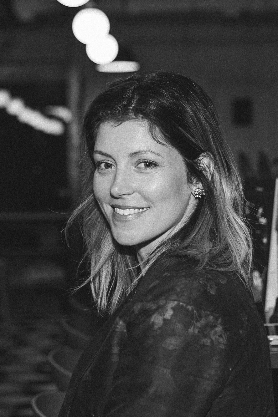 A woman with shoulder-length hair smiling, wearing earrings and a lace top, in a setting with hanging lights.