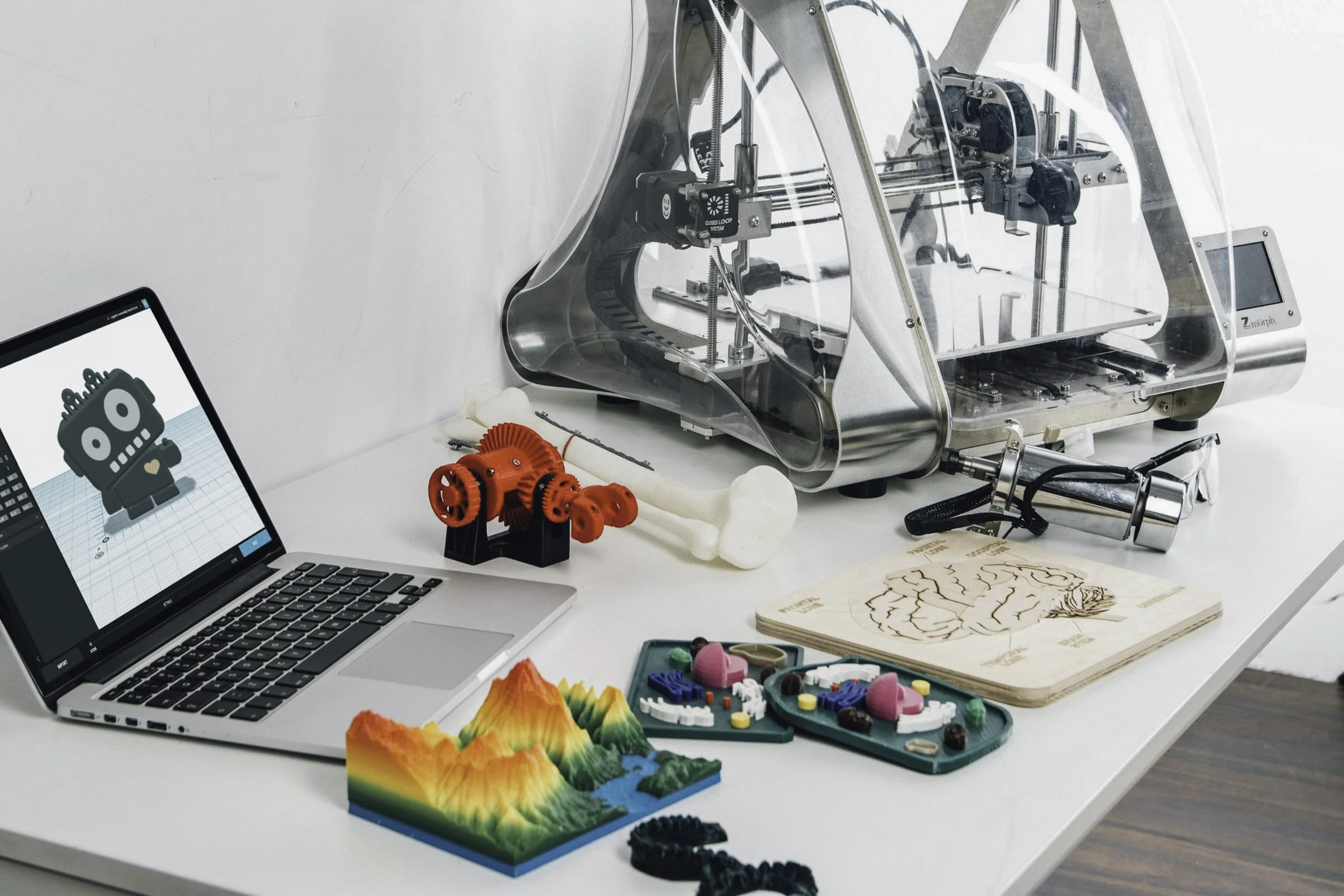 A workspace with a 3D printer, a laptop displaying a robot model, a colorful mountain terrain, a bone with a gear model, and various tools and colorful educational models on a white table.