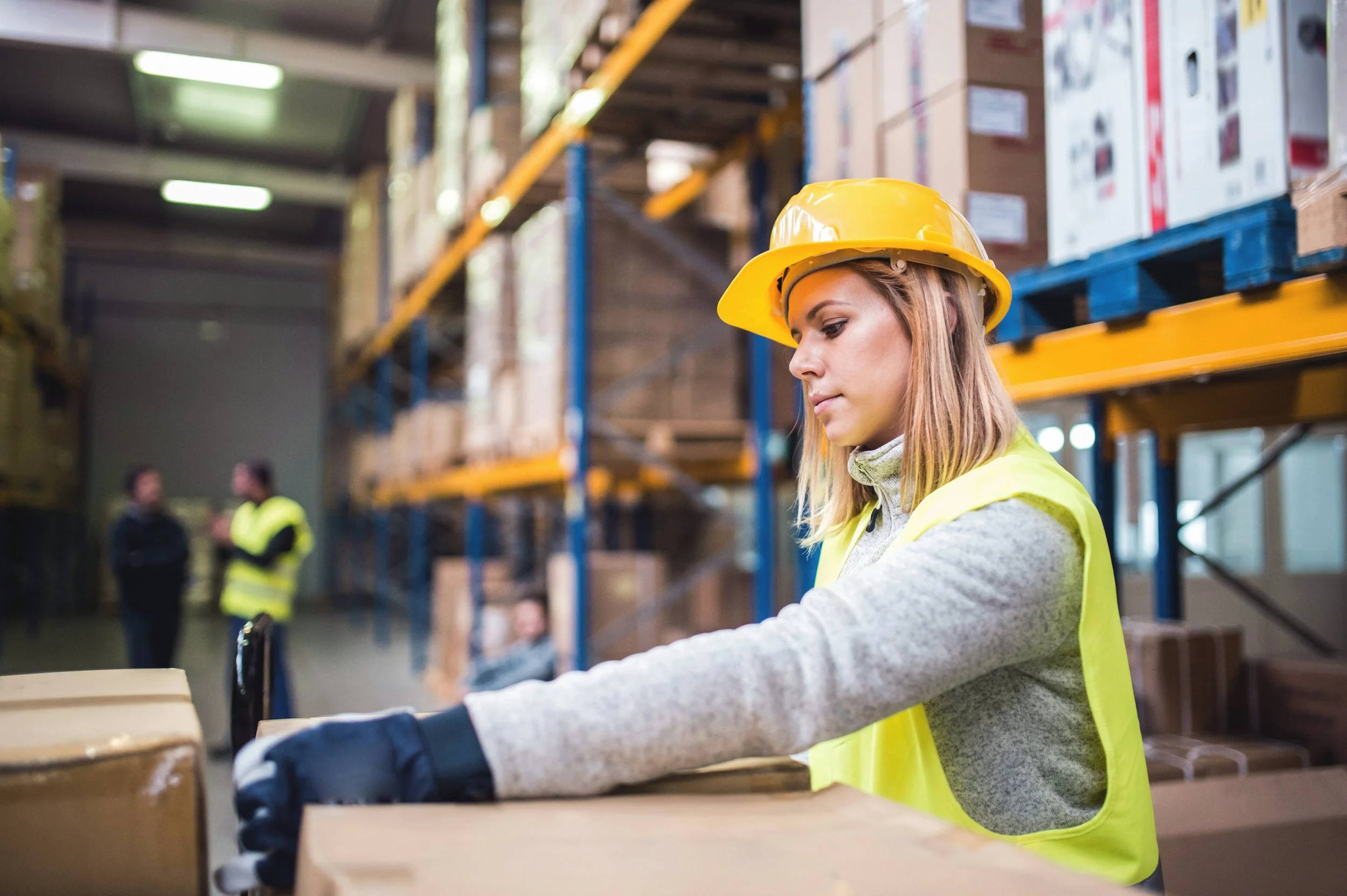 A woman in a yellow safety helmet and vest inspecting boxes in a warehouse with forklifts and shelves in the background.