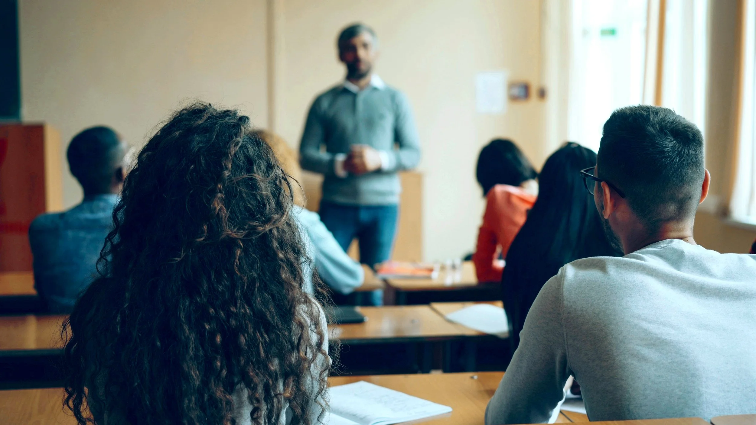 A classroom with a teacher standing at the front and students sitting at desks, some taking notes.