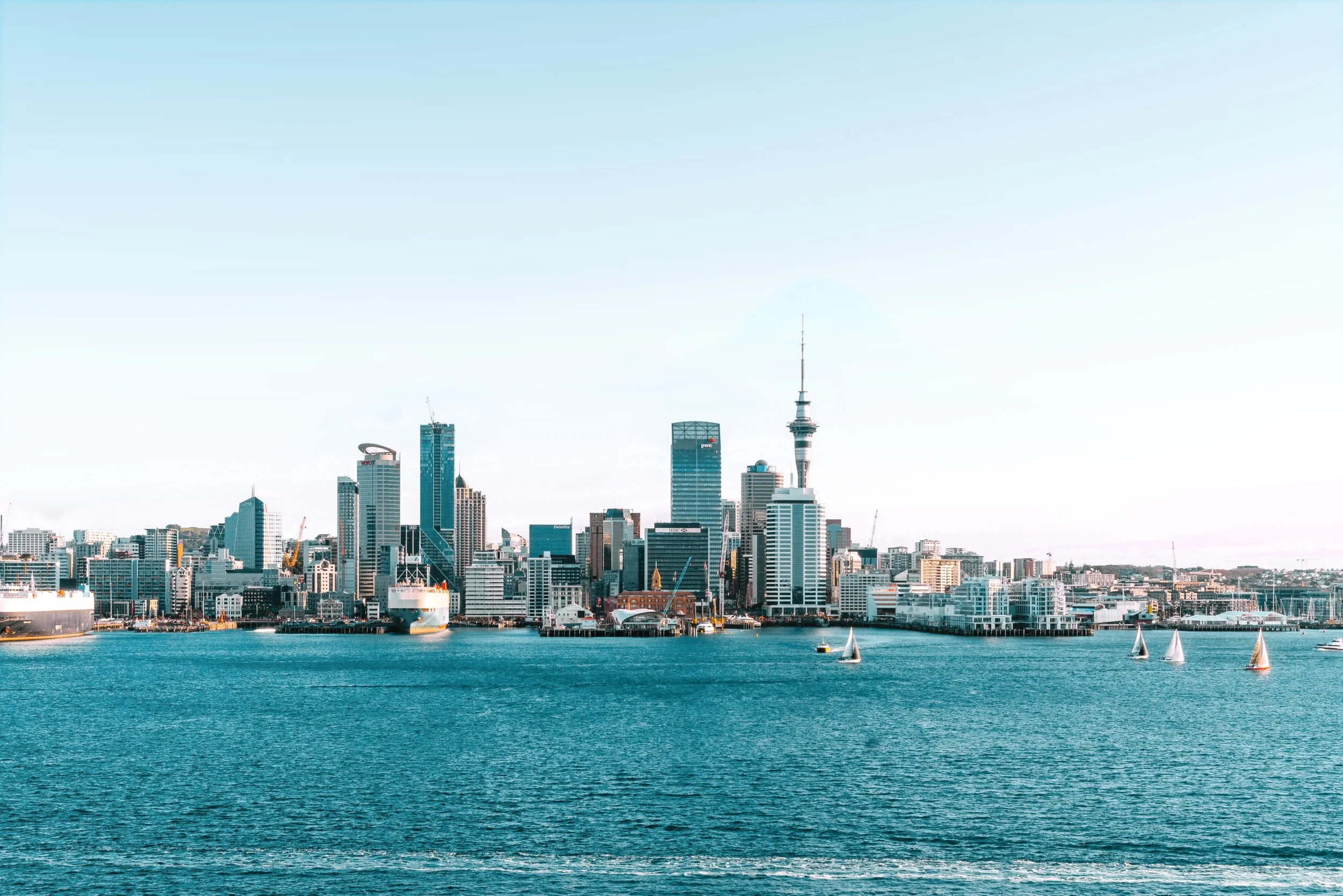 A panoramic view of a city skyline along a waterfront, including high-rise buildings and a prominent tower, with sailboats visible on the water.