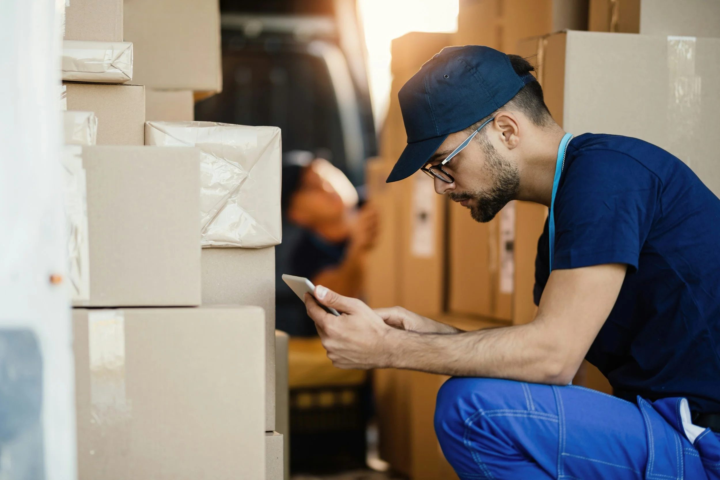 A man wearing glasses, a blue cap, and a navy T-shirt crouches among stacked cardboard boxes, looking at his smartphone.