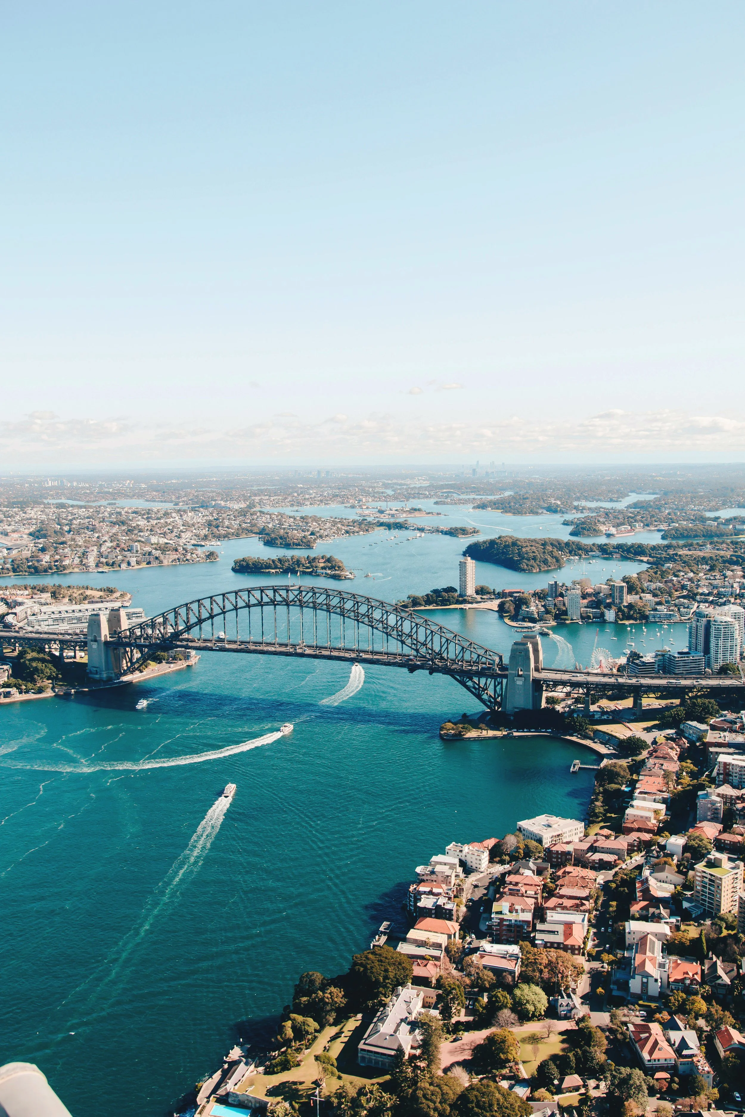 Aerial view of Sydney Harbour Bridge, boats on the water, surrounding cityscape, and distant skyline on a clear day.