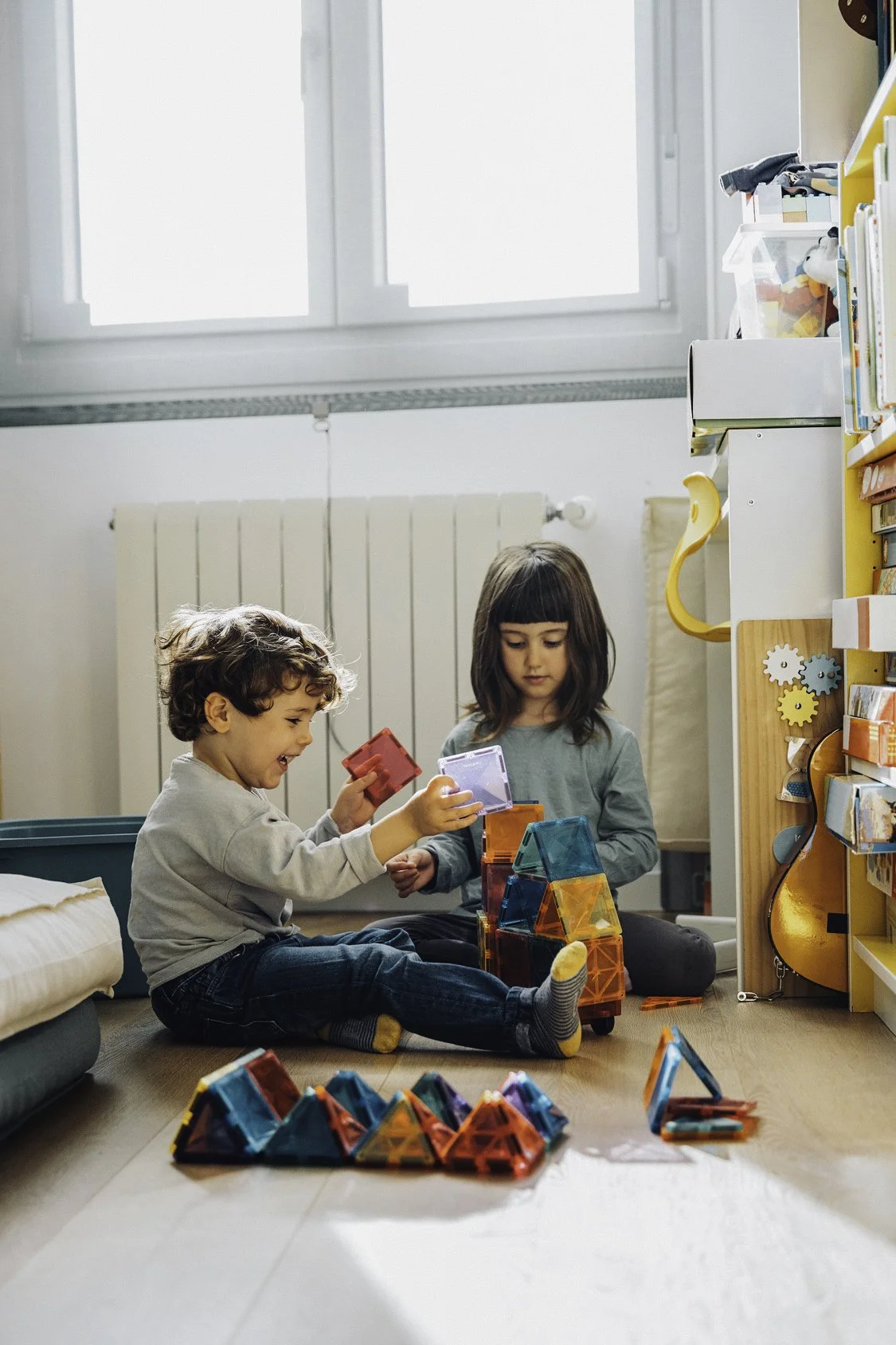 Two children playing with colorful magnetic building tiles on the floor of a brightly lit room, with shelves and a guitar in the background.