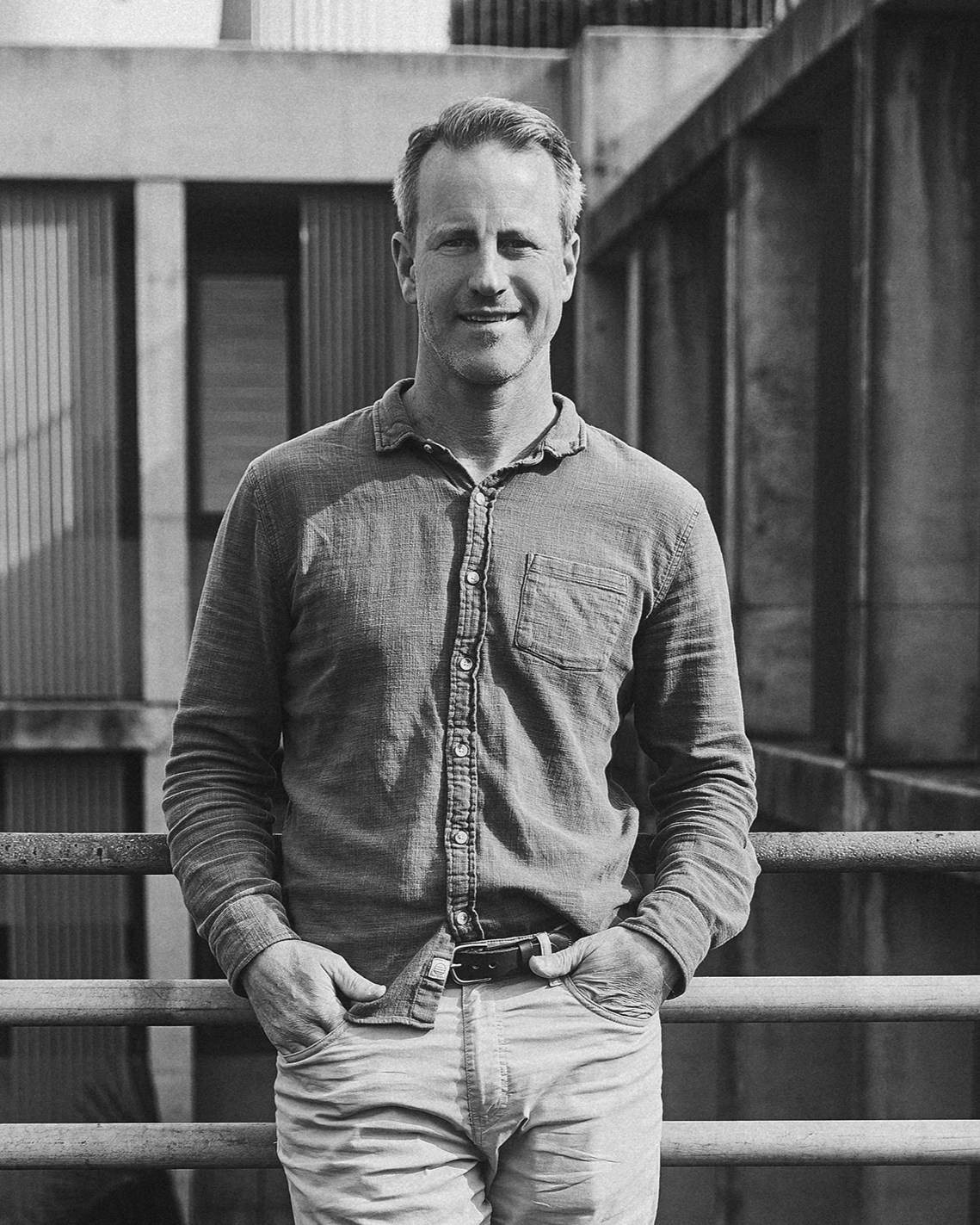 A smiling man in casual clothes standing outdoors, leaning on a railing with modern building architecture in the background.