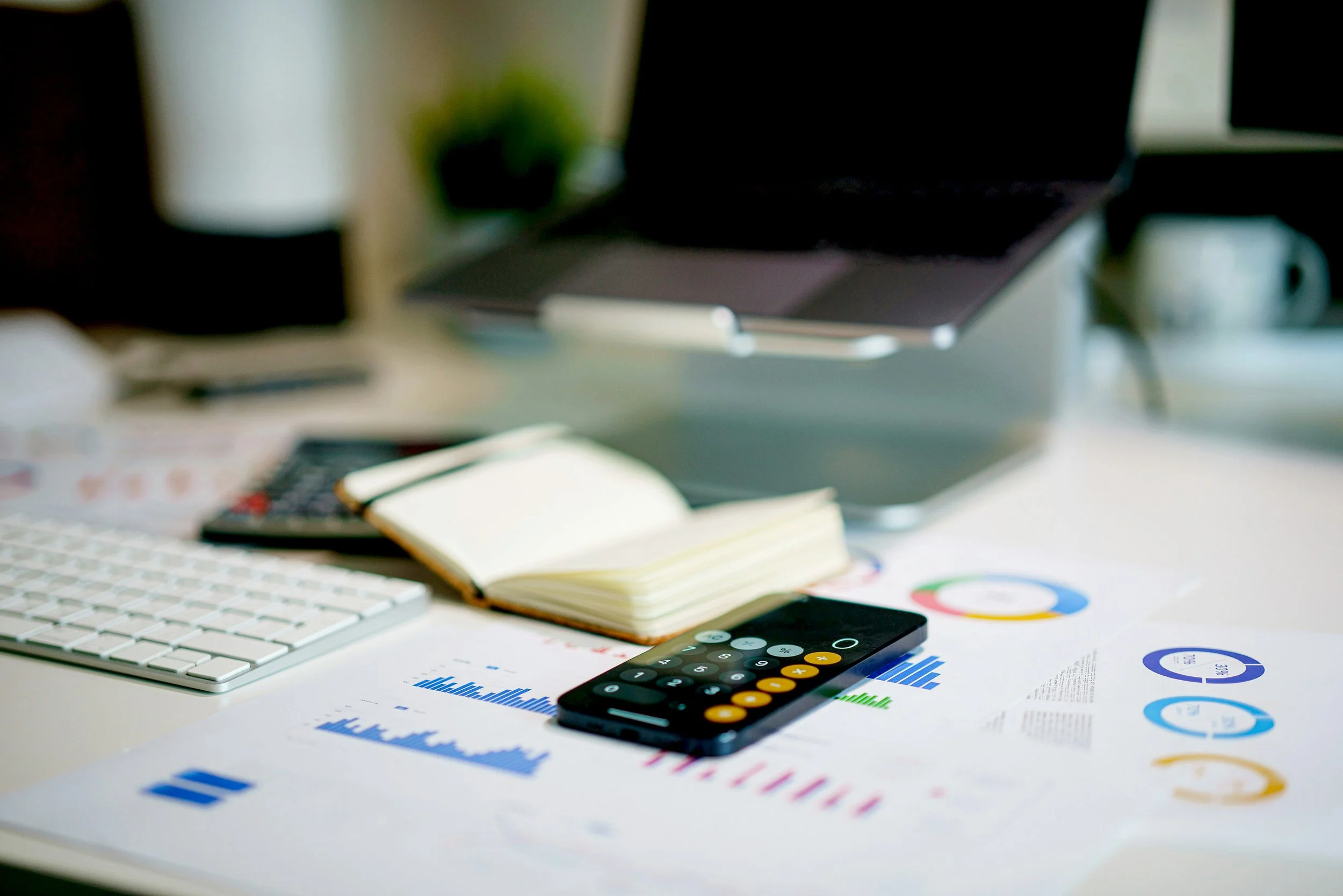 Desk with financial charts, calculator, keyboard, open notebook, pen, laptop, and coffee mug.