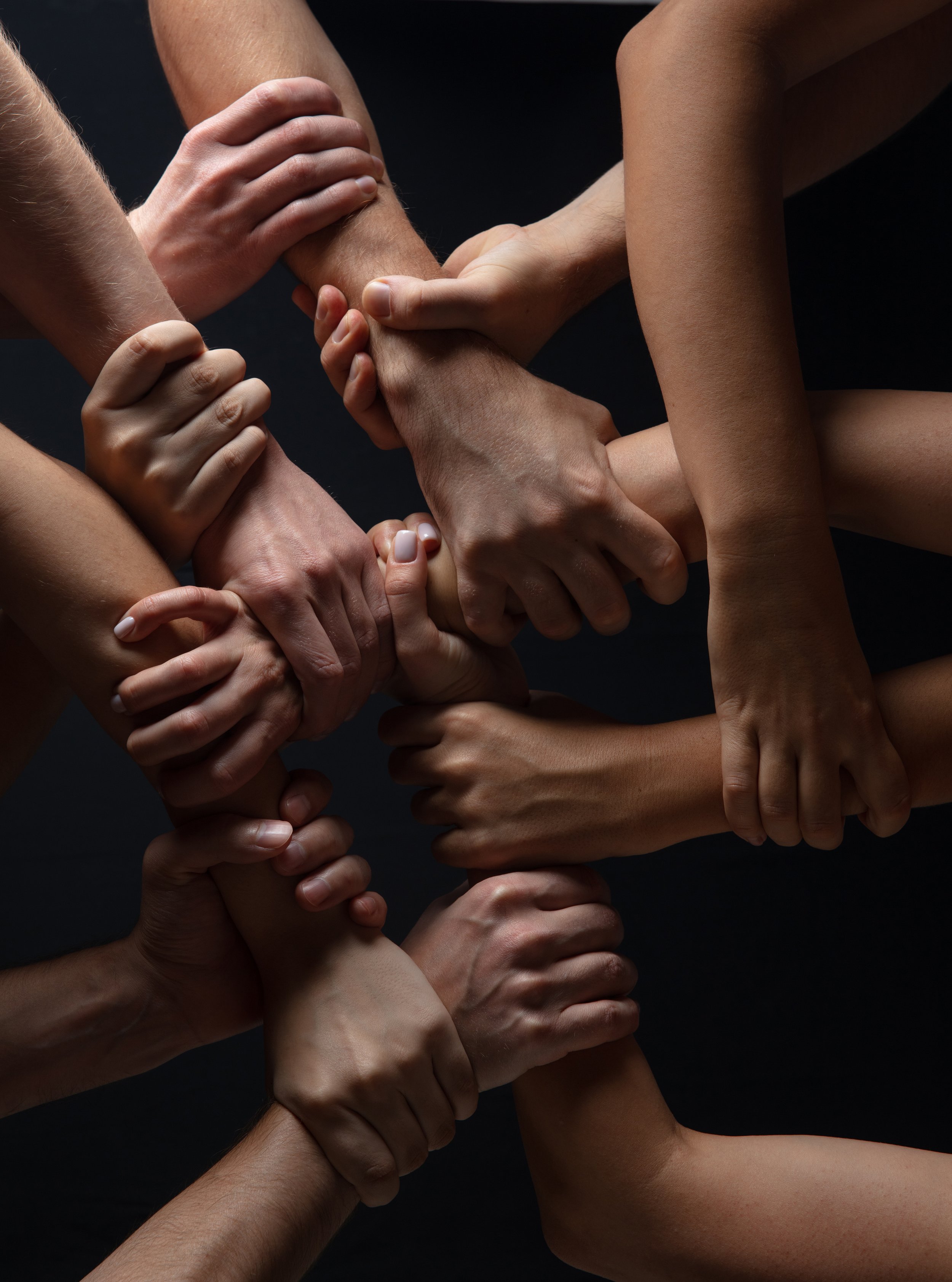 Multiple hands of diverse skin tones gripping each other's arms in a circle against a dark background.