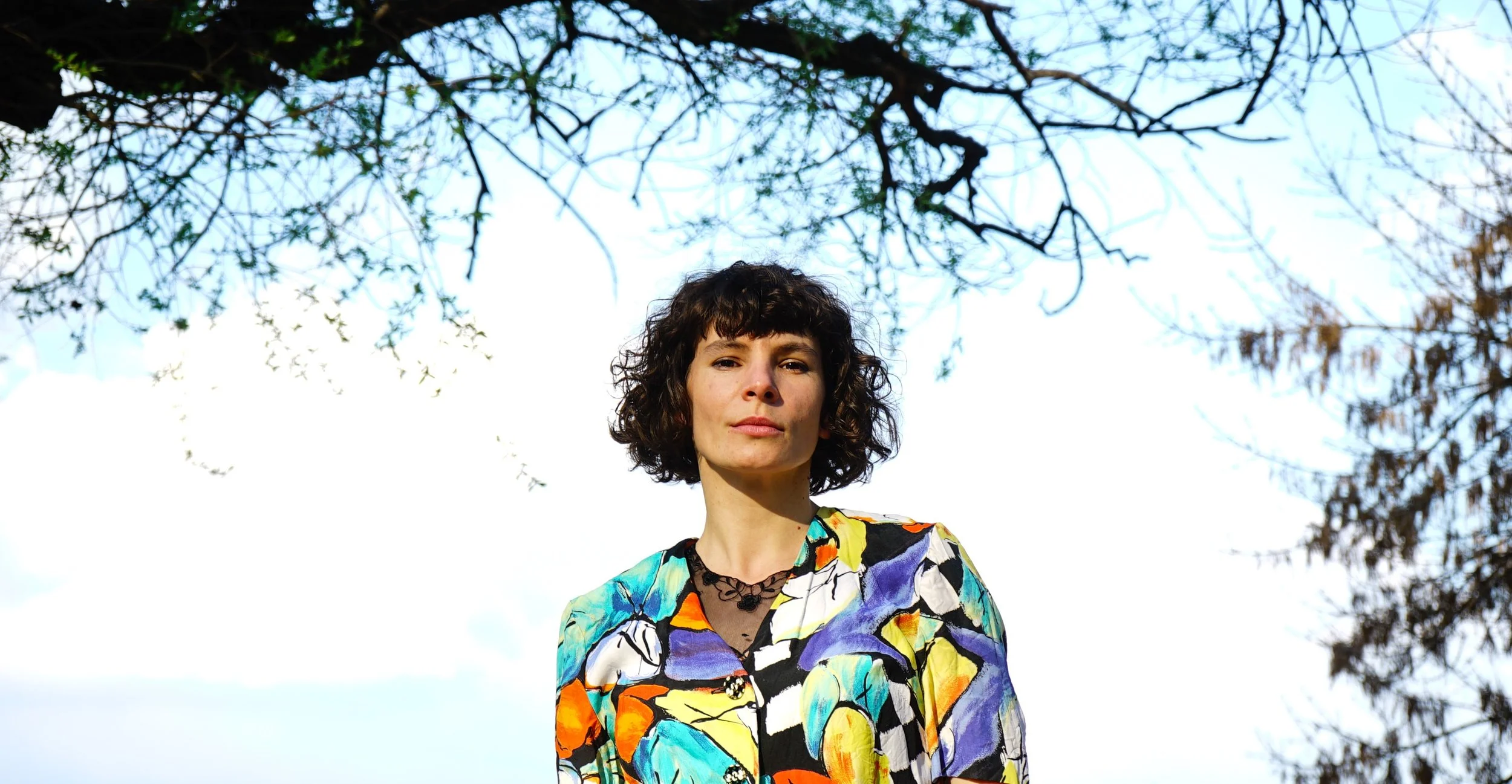 A woman with curly dark hair wearing a colorful, patterned shirt stands outdoors beneath a tree with sparse leaves, against a bright sky.