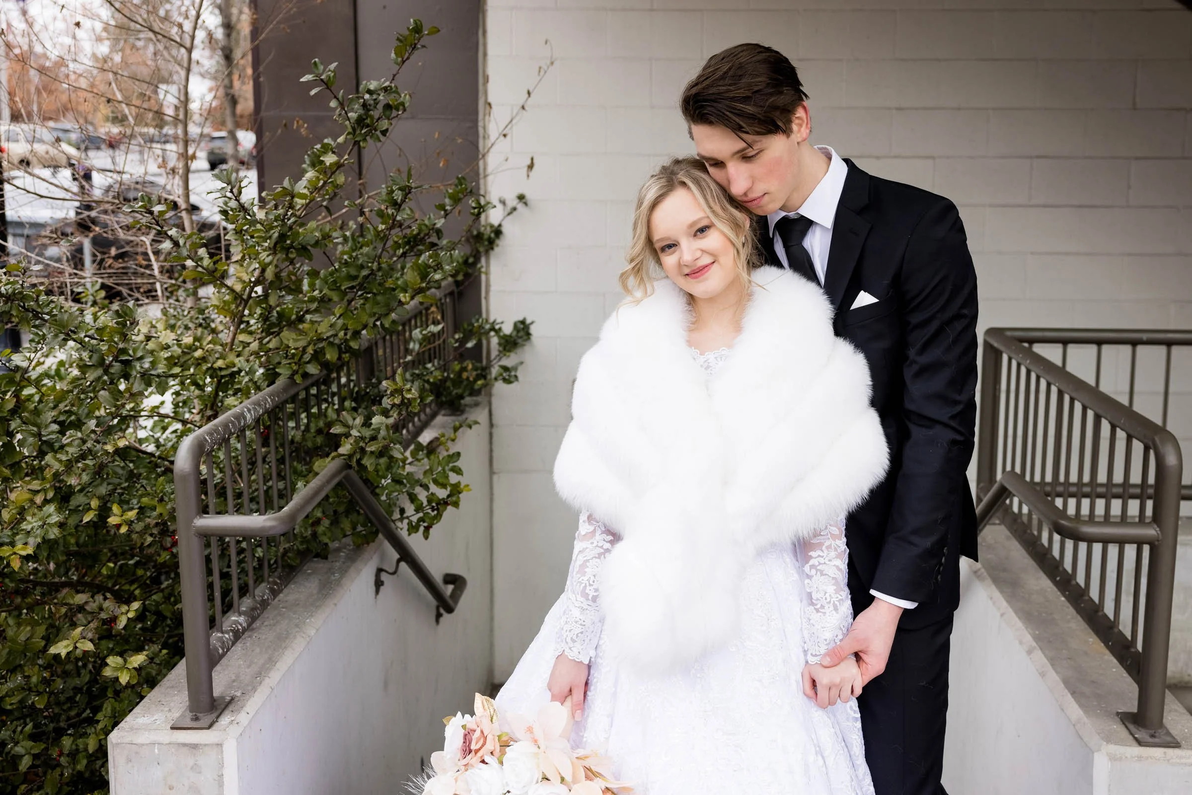A young couple dressed in wedding attire, standing outdoors on a staircase, holding hands, with the woman holding a bouquet of flowers, and the man leaning on her shoulder. Spokane wedding photos.