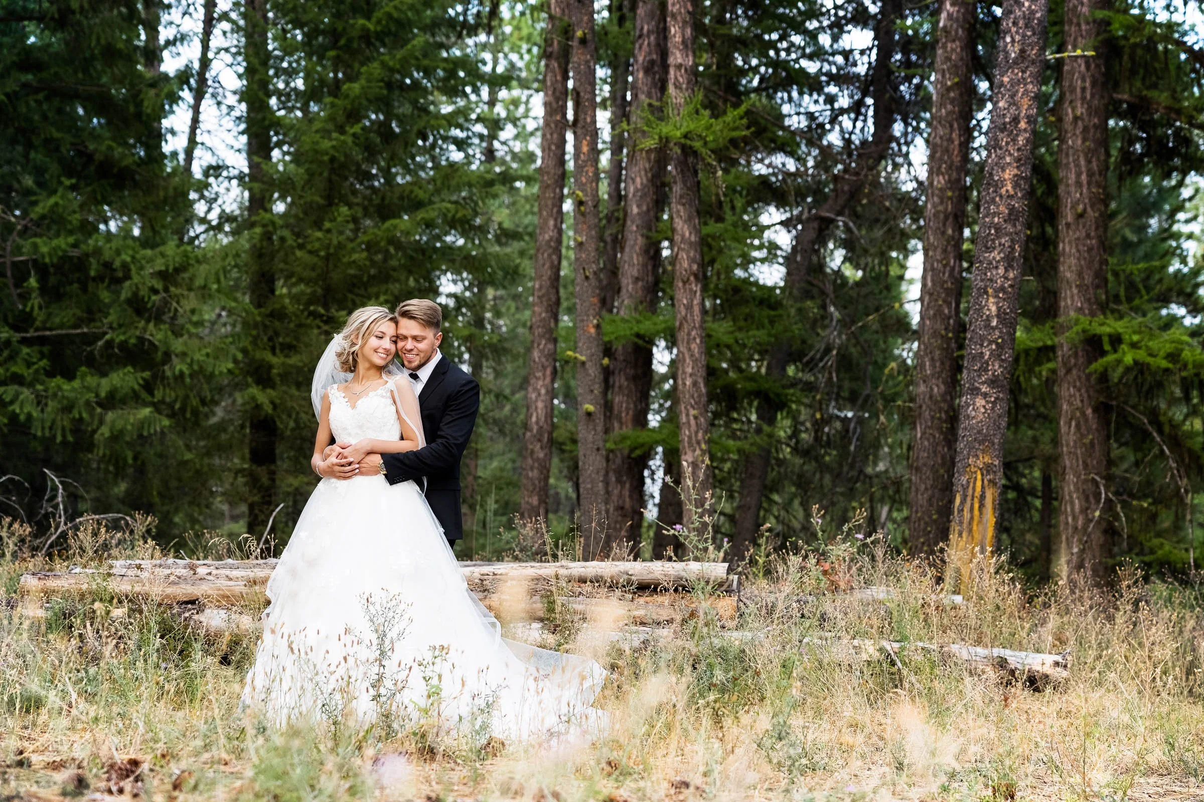 A bride and groom are smiling and hugging in a forest clearing with tall trees and green foliage. Spokane wedding photography.