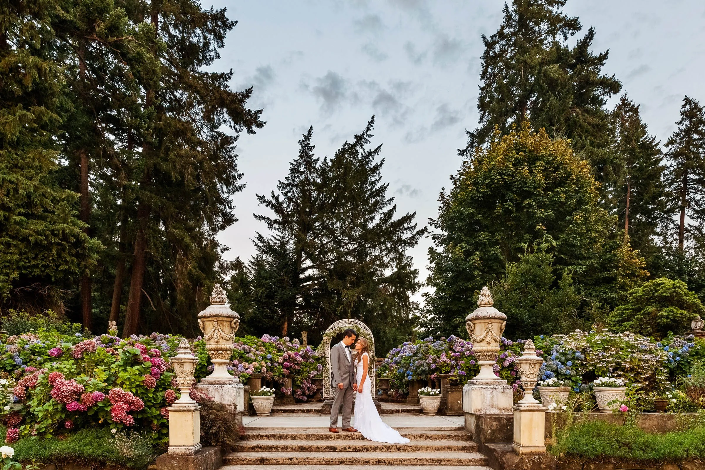 A couple dressed in wedding attire standing on steps in front of an archway, surrounded by blooming hydrangea bushes and stone urns, with tall trees and a partly cloudy sky in the background.