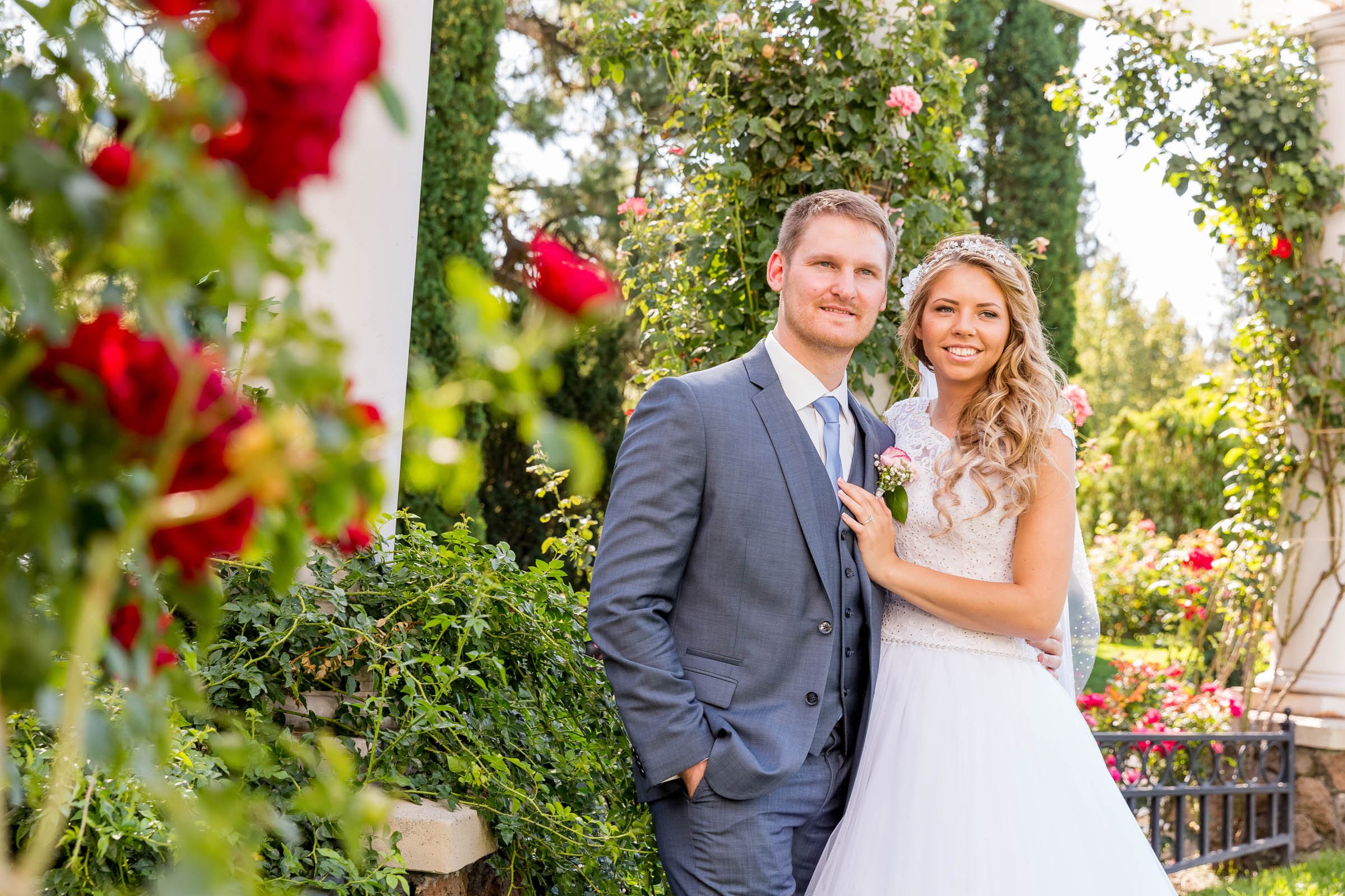 A newlywed couple standing outdoors among greenery and pink flowers, smiling for a photo. The groom is wearing a gray suit with a tie, and the bride is in a white wedding dress with lace details, with her arm around the groom's shoulder.